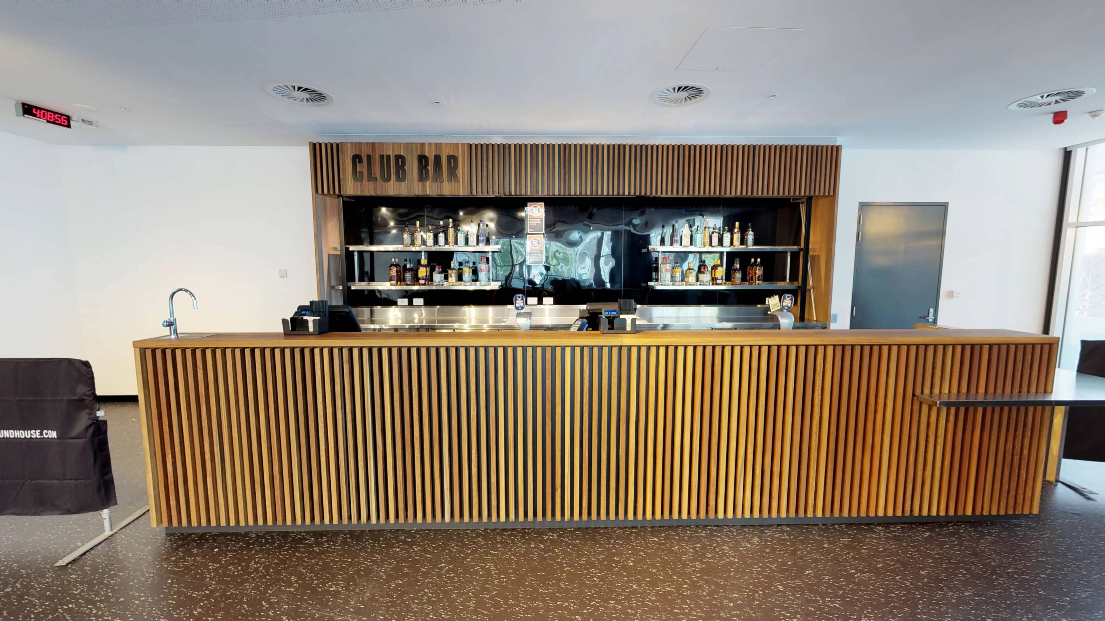 A modern bar area with a wooden slatted counter, shelves stocked with various bottles, and a minimalist design.