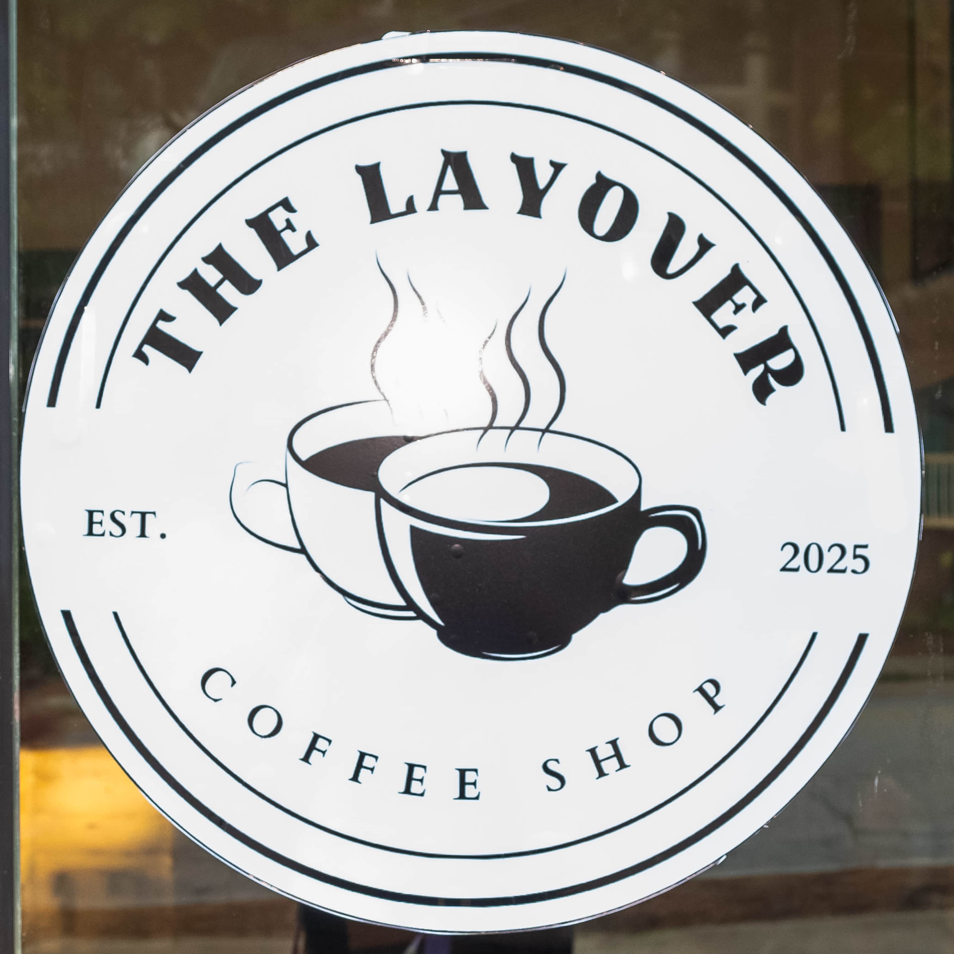 The image shows a circular logo for "The Layover Coffee Shop," featuring two steaming coffee cups and the establishment year "2025."