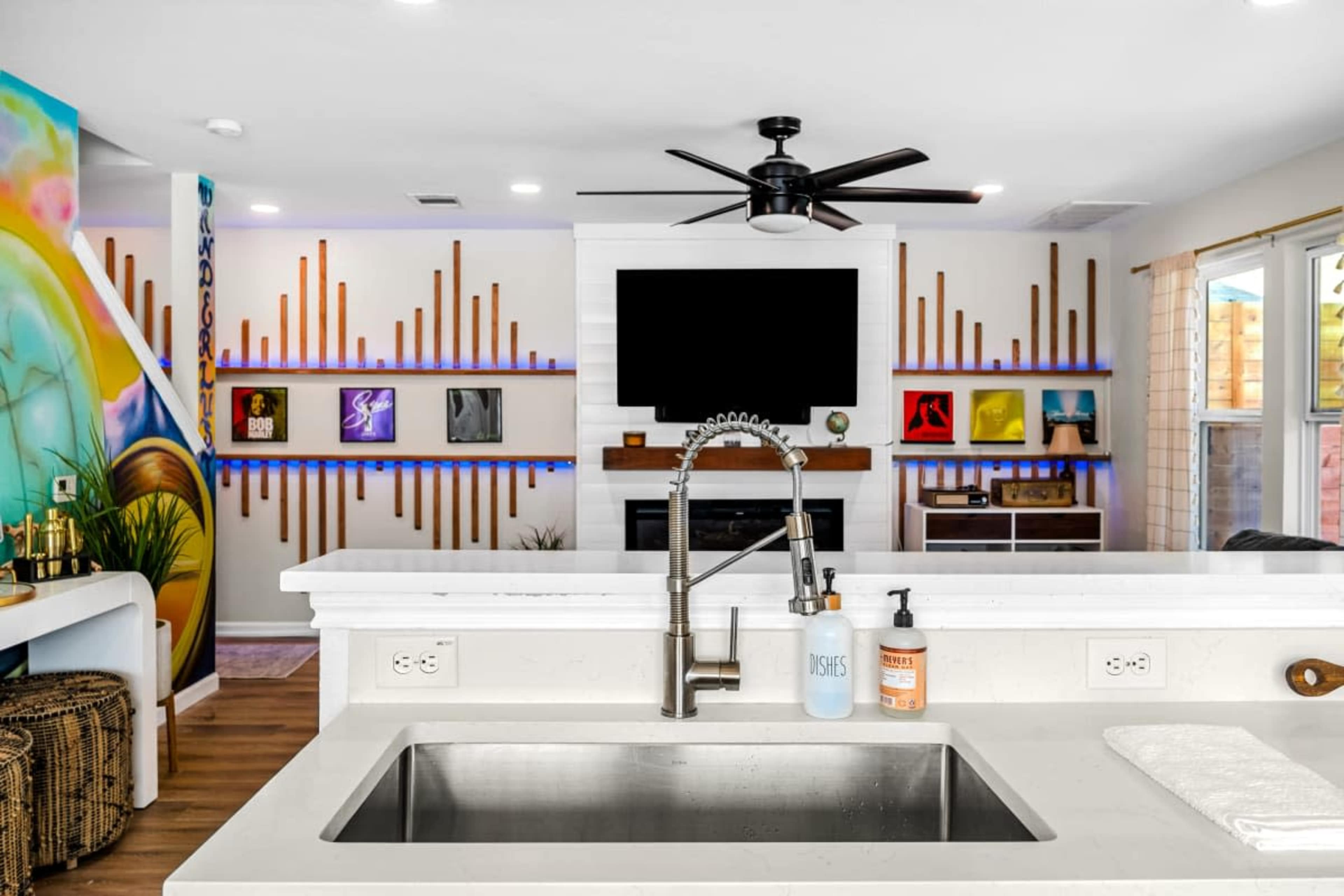 A modern kitchen and living area with a large sink, a stylish ceiling fan, and a wall-mounted television above a decorative fireplace.