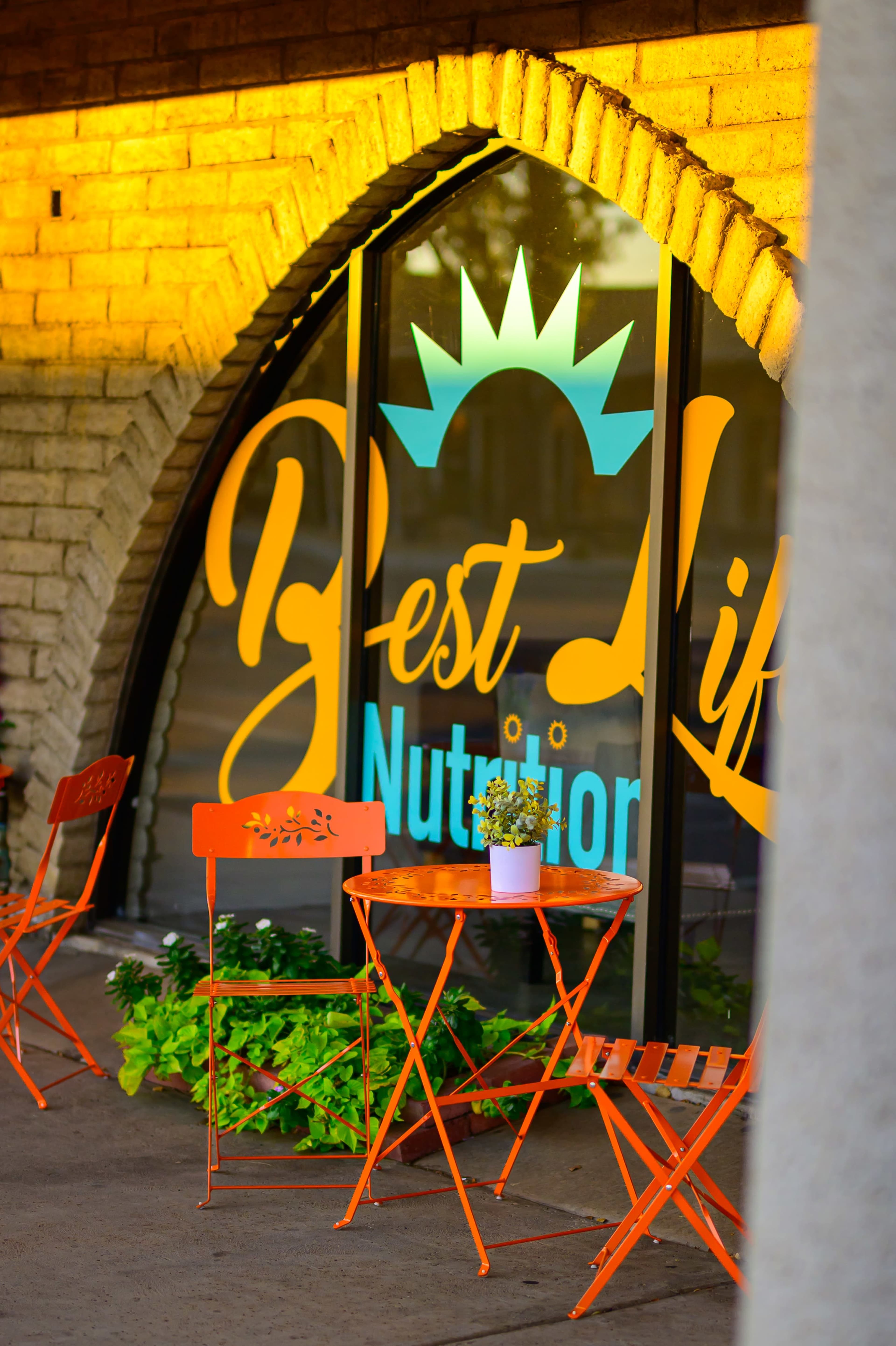 The image shows a small outdoor seating area with orange chairs and a table in front of a shop window that displays the words "Best Life Nutrition."