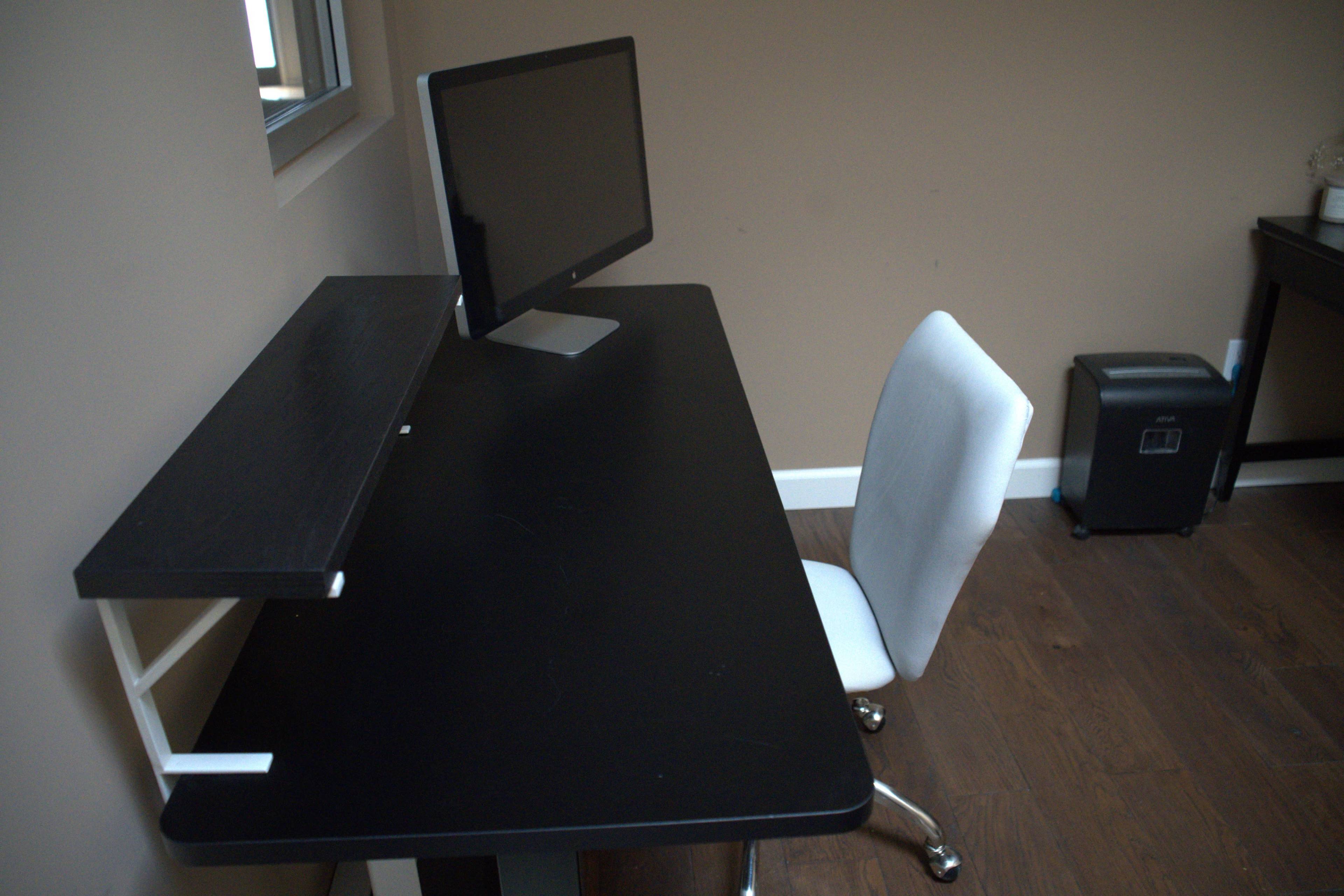 The image shows a simple black desk with a raised shelf, a computer monitor, and an empty white chair in a room with brown walls and wooden flooring.