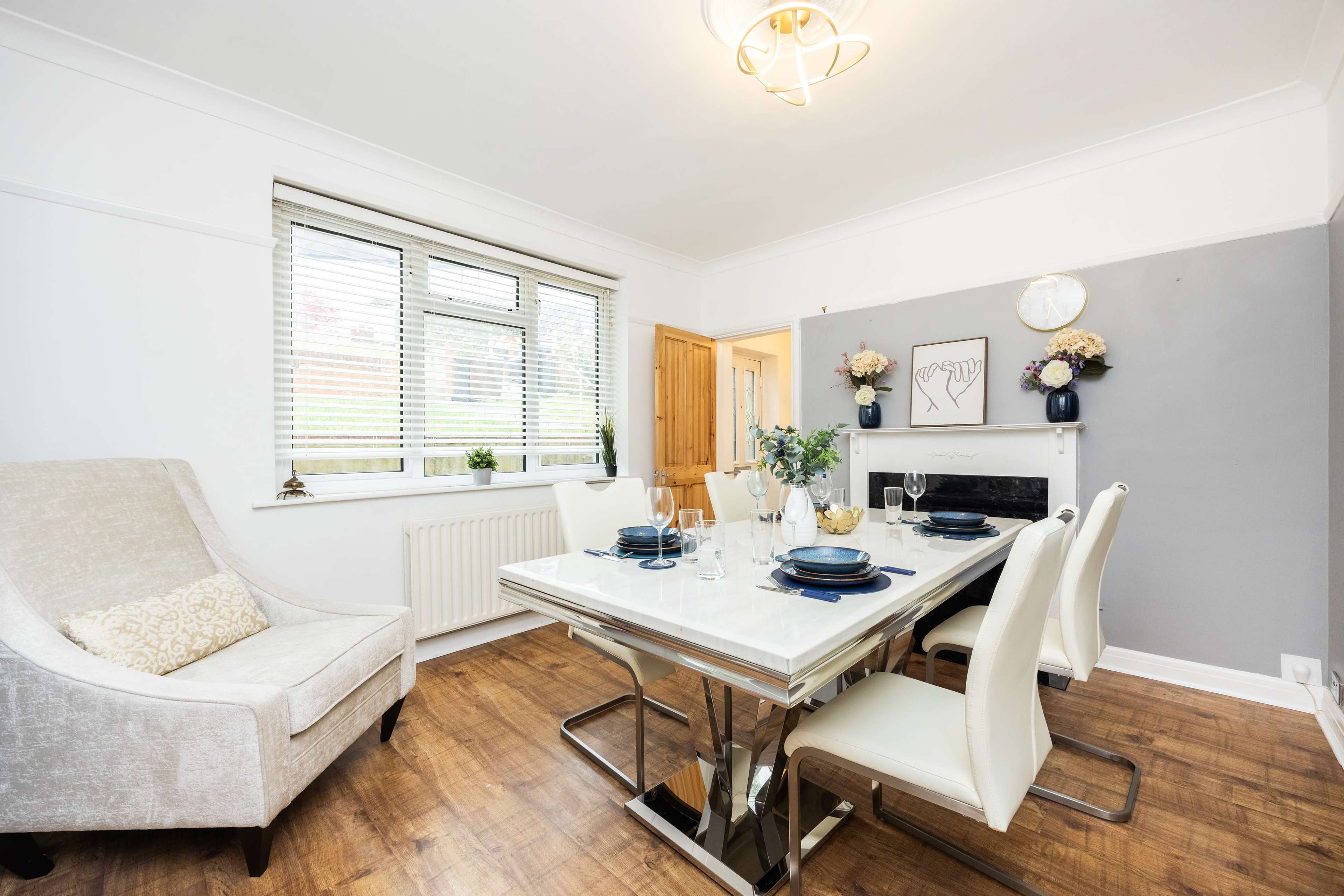 A modern dining room features a sleek table set for dinner, with an elegant light fixture overhead and a cozy armchair in the corner.
