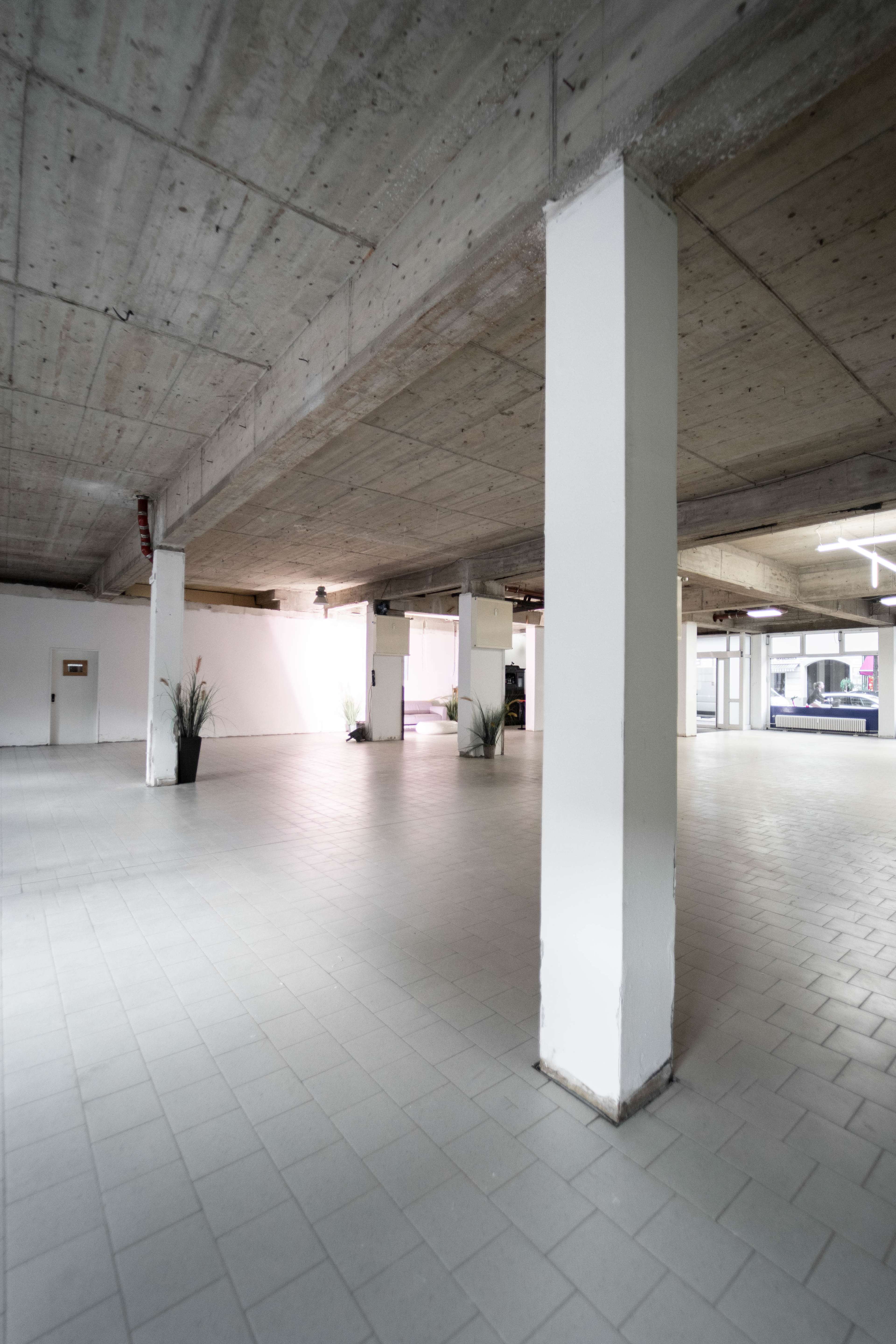 A spacious, empty indoor area with concrete walls and columns, featuring tiled flooring and minimal furnishings.