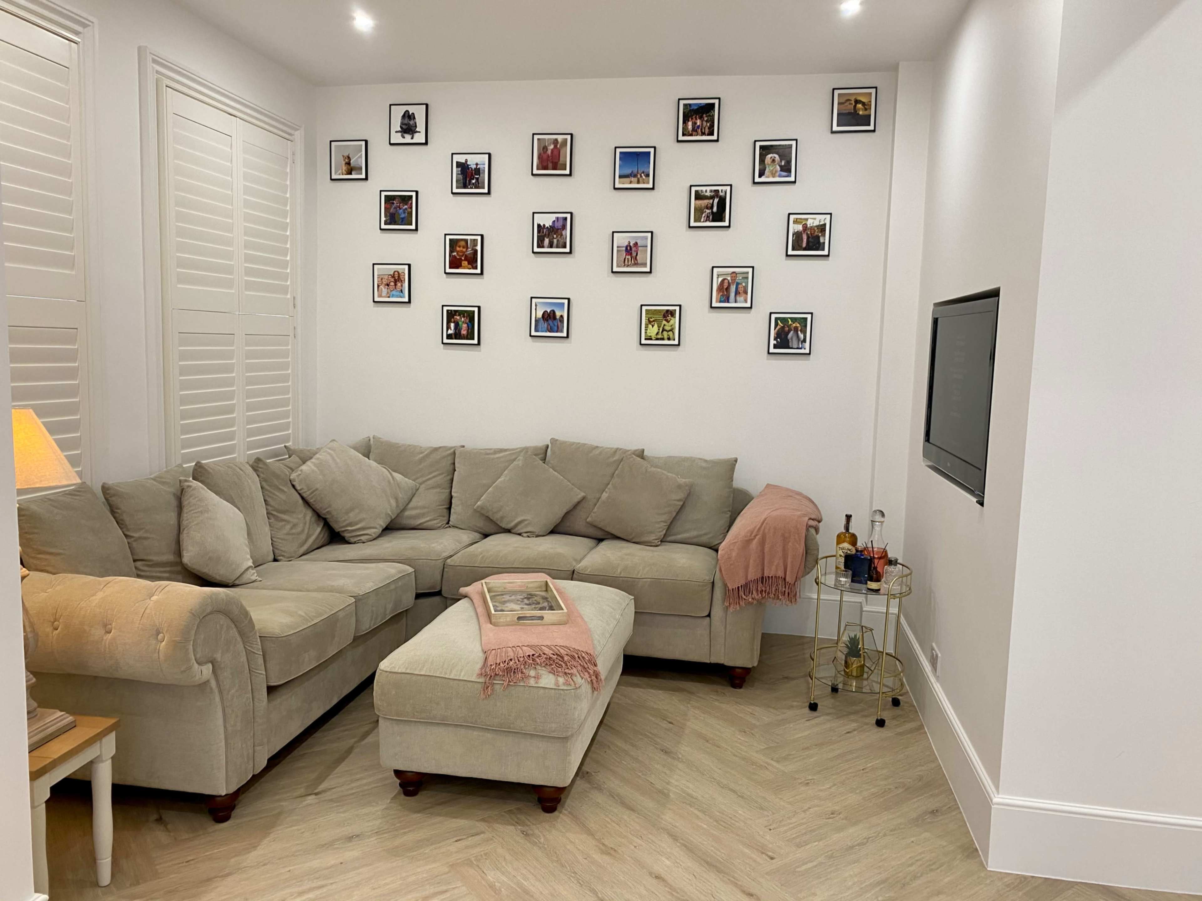 A cozy living room features a sectional couch with decorative pillows, a light blanket draped over the arm, a small ottoman, and a wall adorned with framed photos.