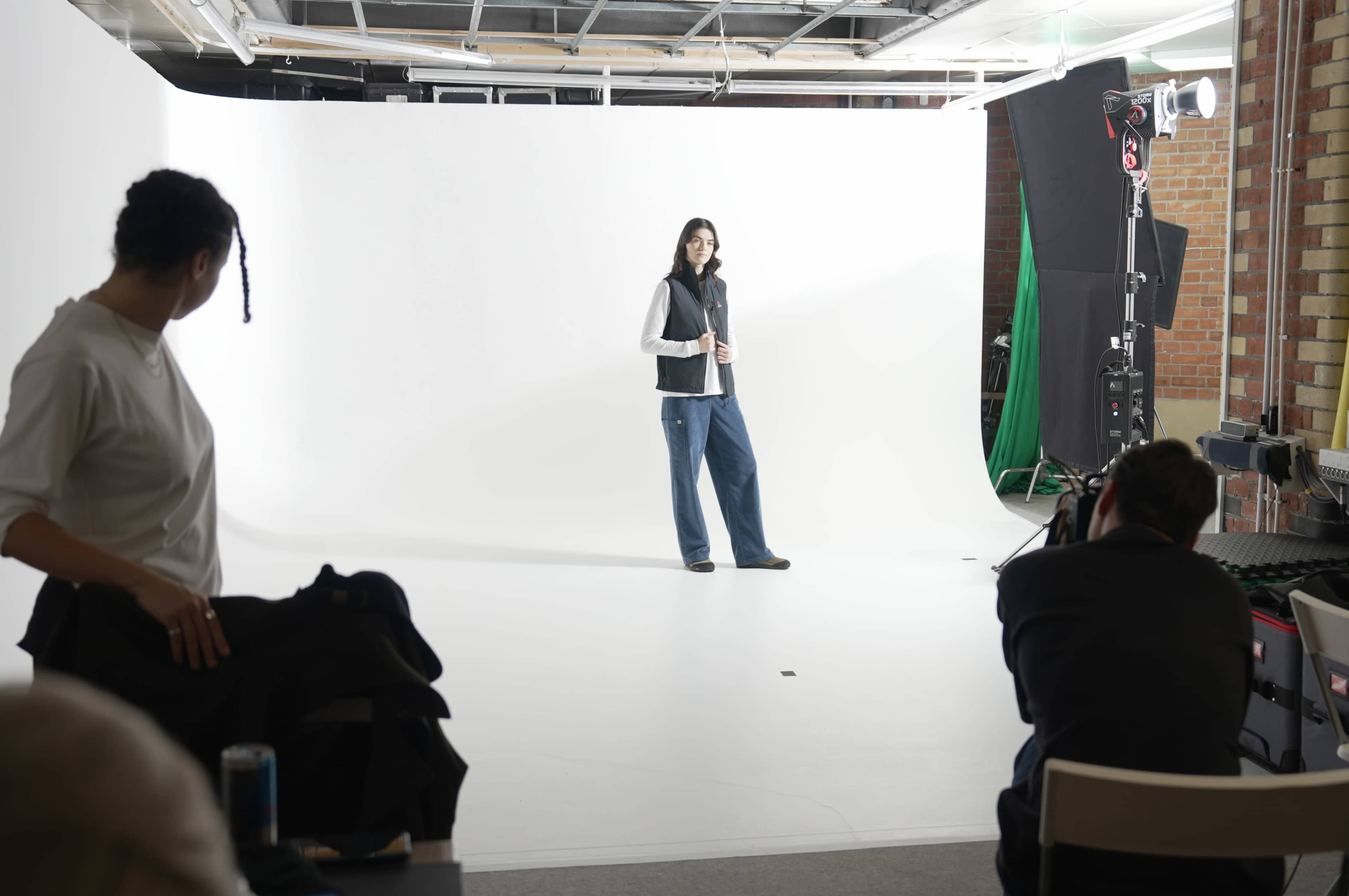 A model stands on a white backdrop while a photographer takes pictures, and another person observes in a studio.