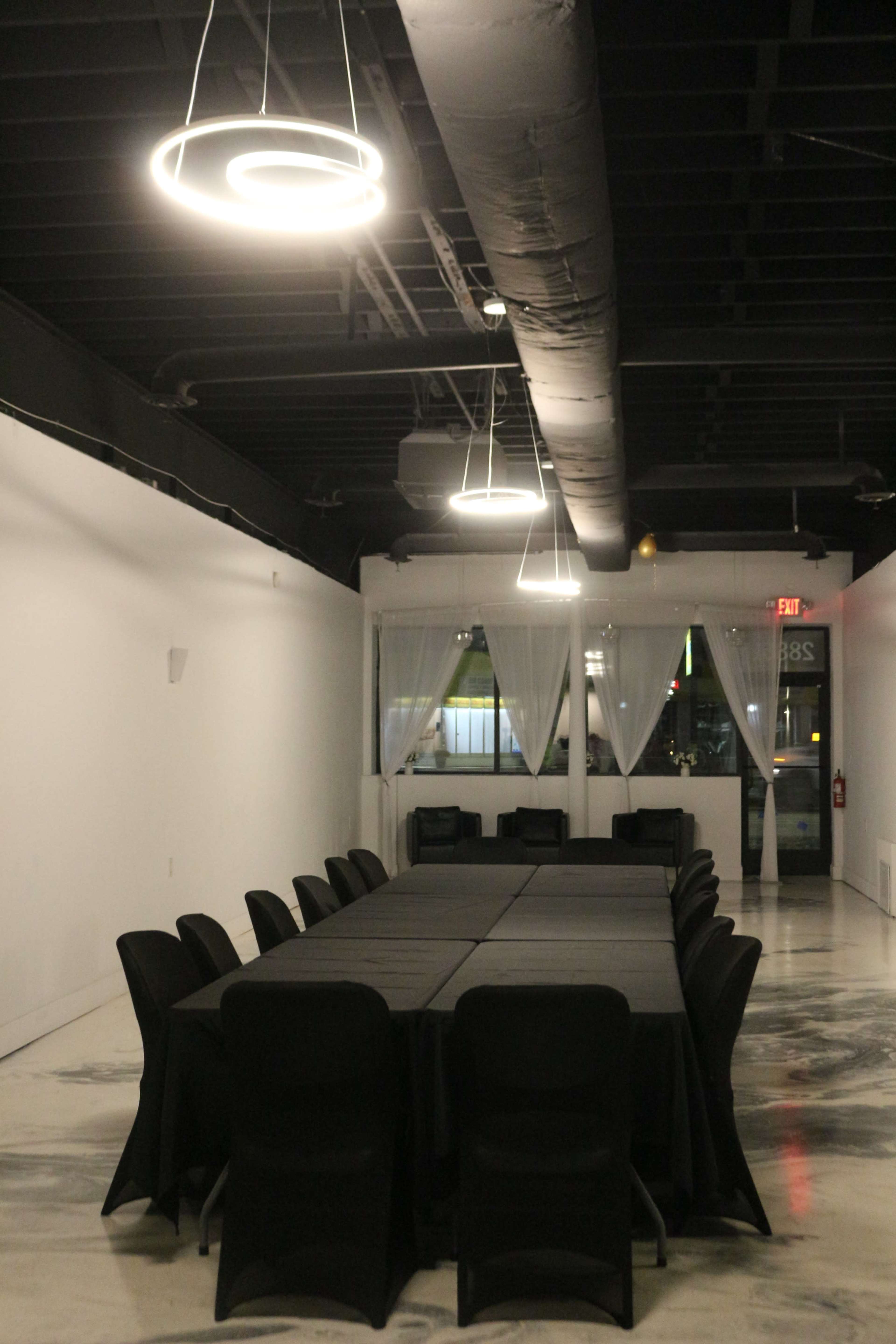 A long, rectangular table with black tablecloths is set in a dimly lit meeting room, surrounded by black chairs beneath modern circular light fixtures.