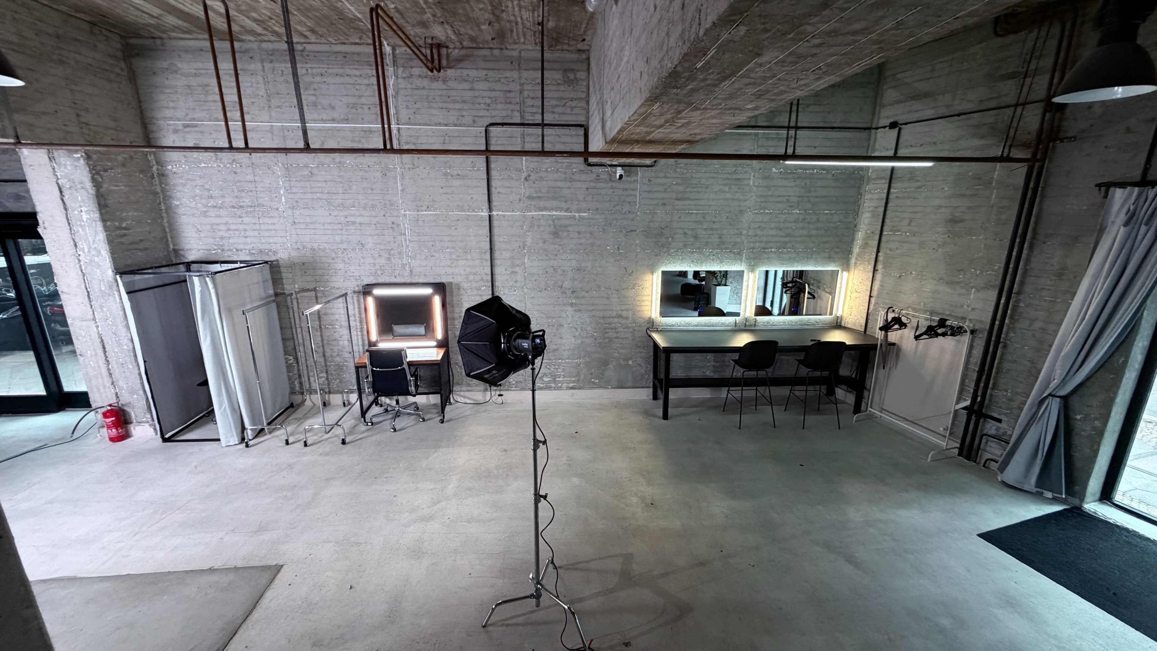 The interior of a spacious studio features exposed brick walls, a large work table with illuminated mirrors, a backdrop stand, and a professional lighting setup.