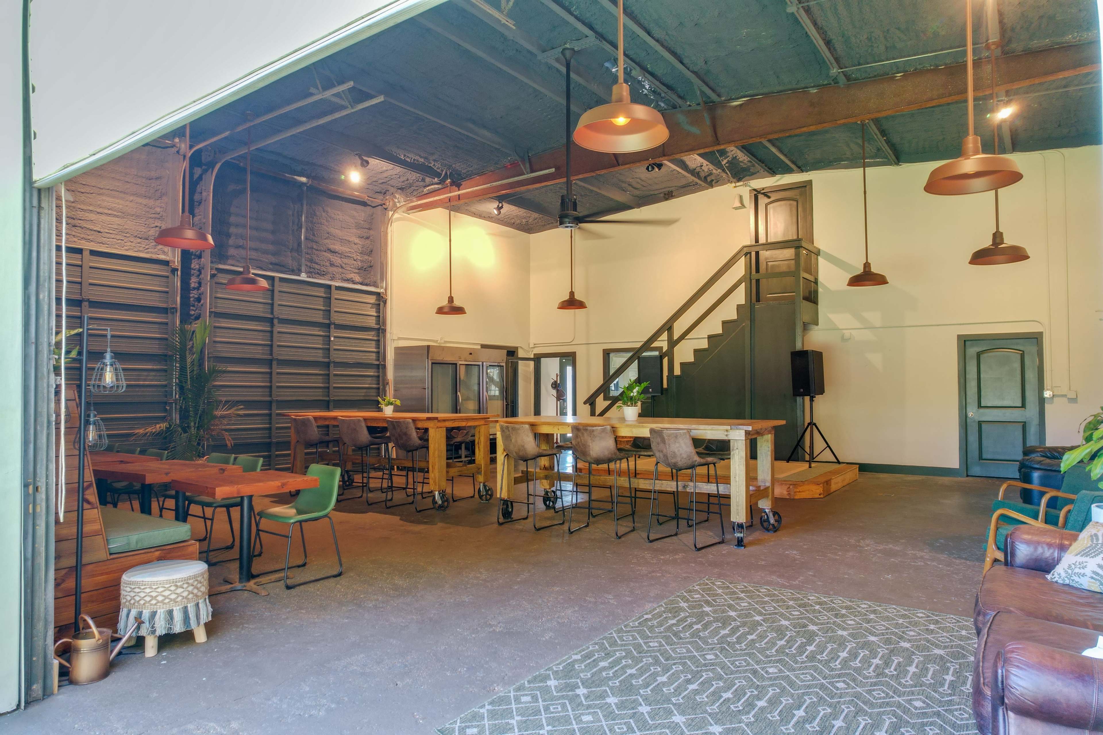 The image shows an open industrial-style space featuring wooden tables, bar stools, and a staircase, with ample natural light illuminating the area.