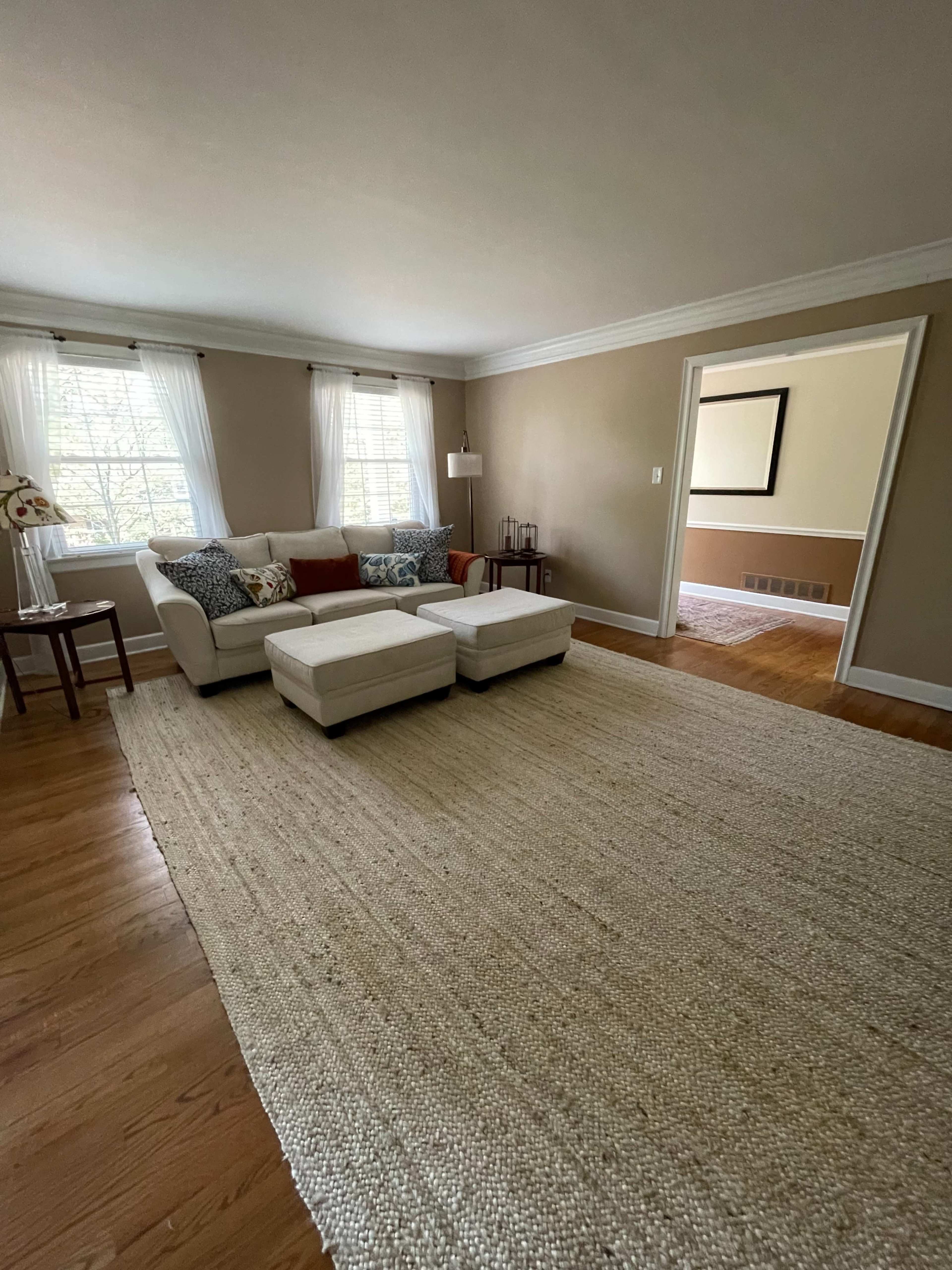 The living room features a light-colored sofa with decorative cushions, two ottomans, a large woven area rug, and windows allowing natural light to enter.