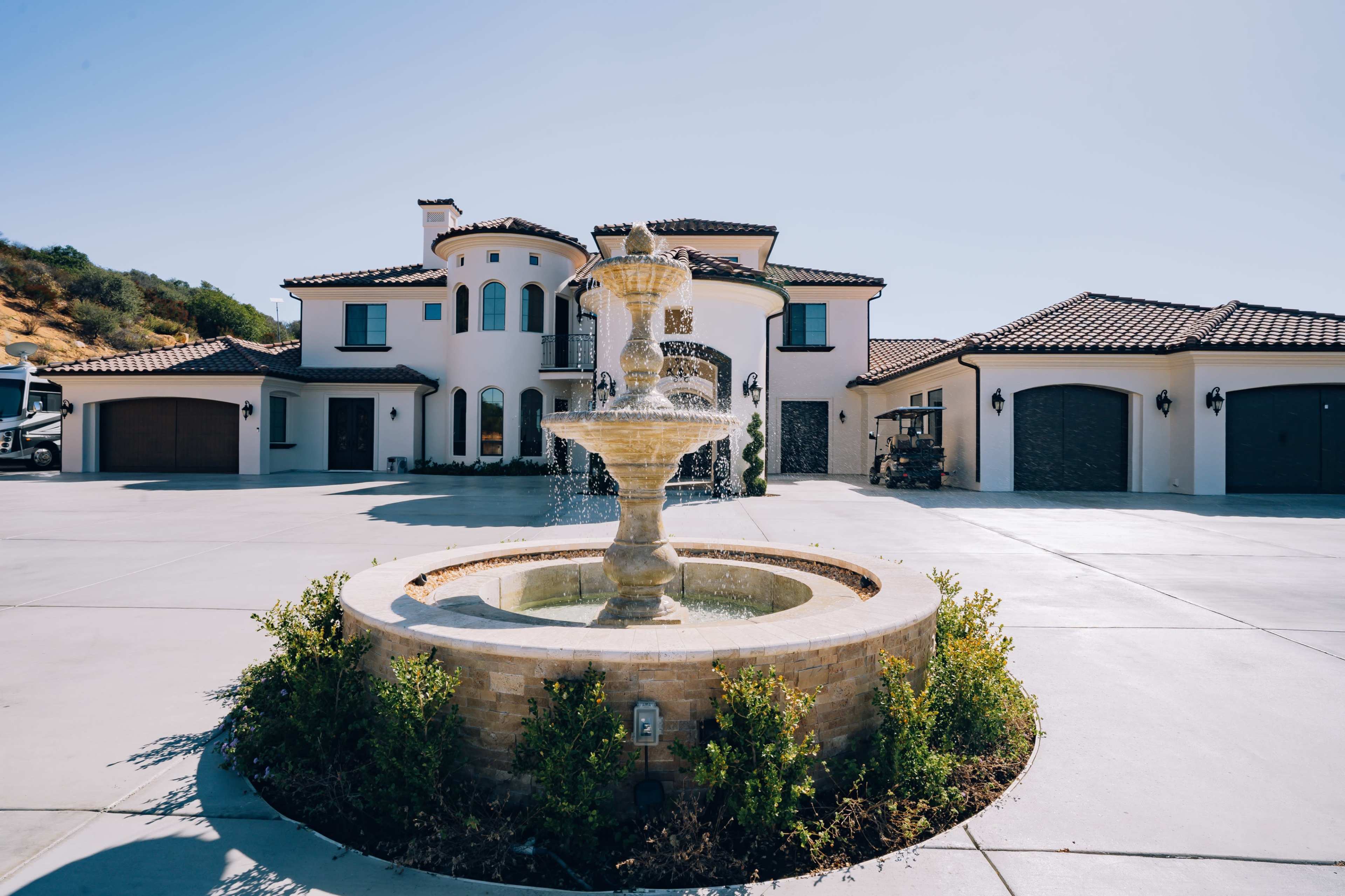 A large fountain is situated in the center of a spacious driveway in front of a modern two-story house with a Mediterranean architectural style.