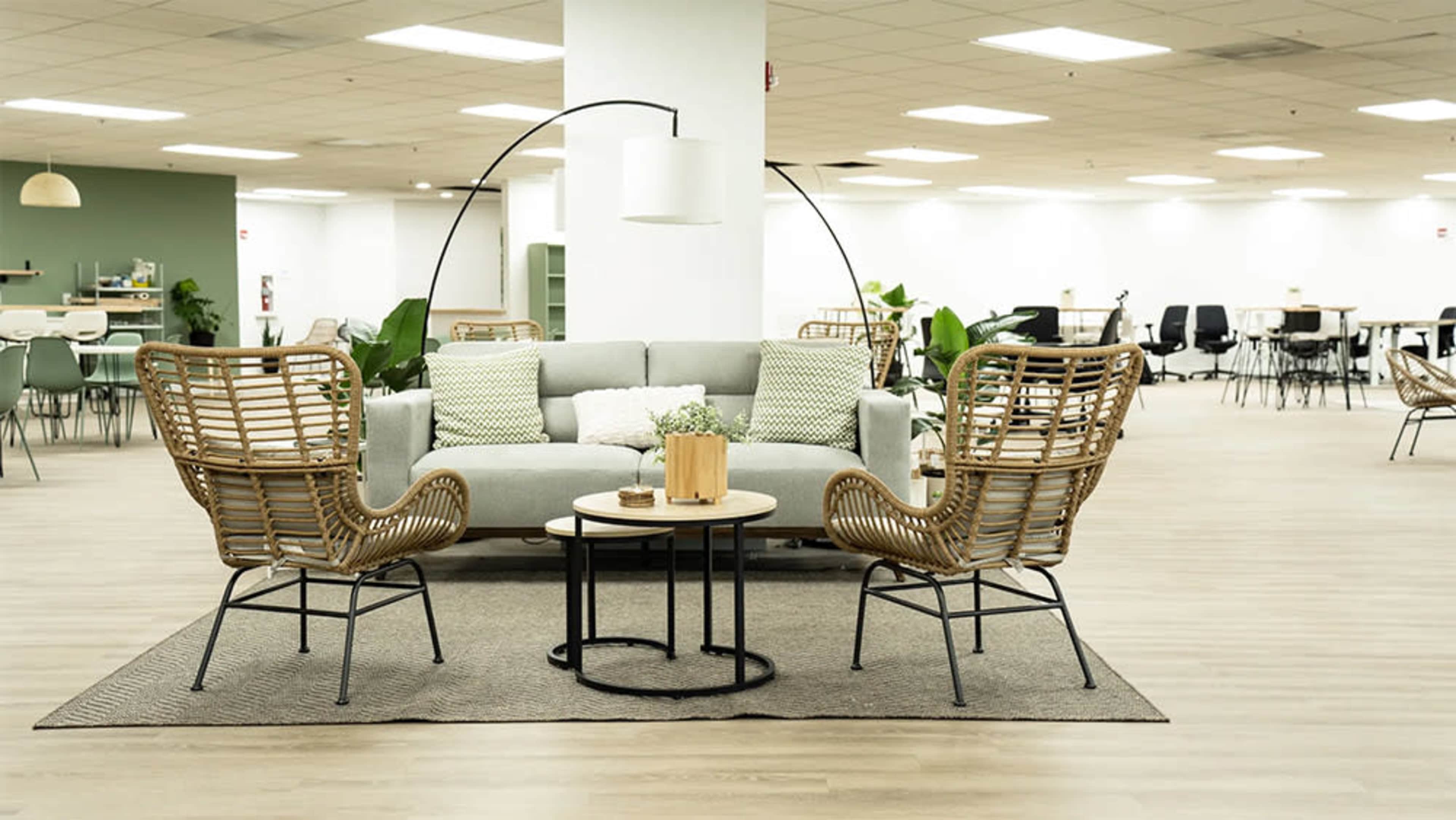 A modern seating area with a gray sofa and two woven chairs arranged around a small circular table on a rug in a spacious, well-lit room.