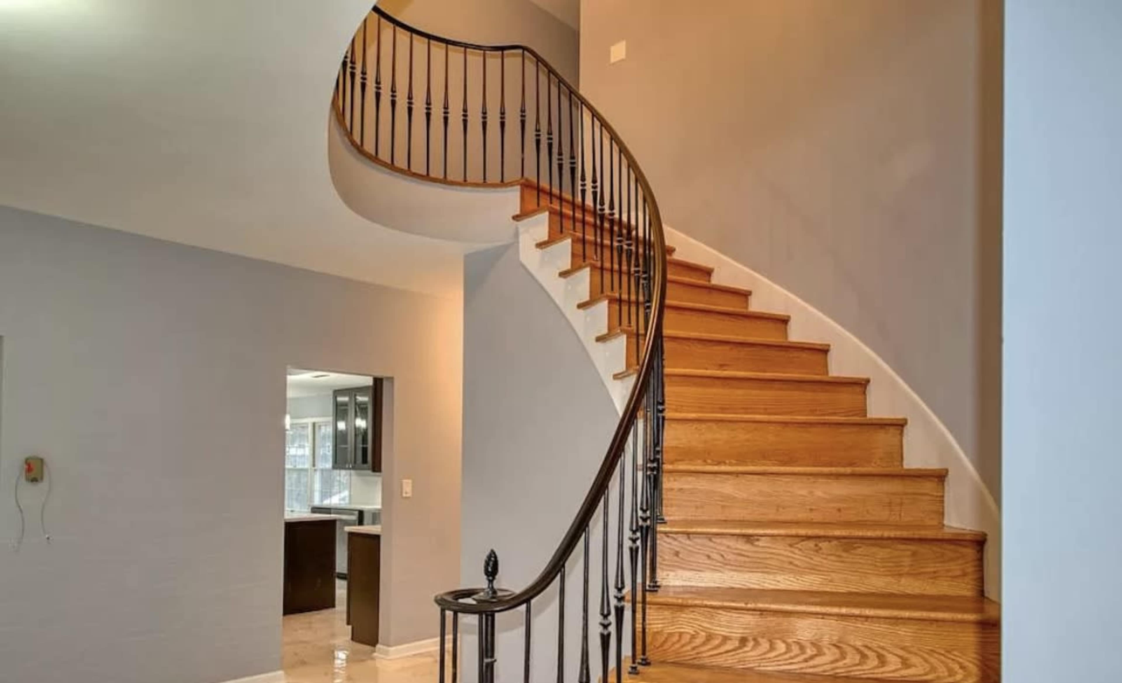 The image shows a wooden spiral staircase with a black wrought iron railing in a light-colored interior space.