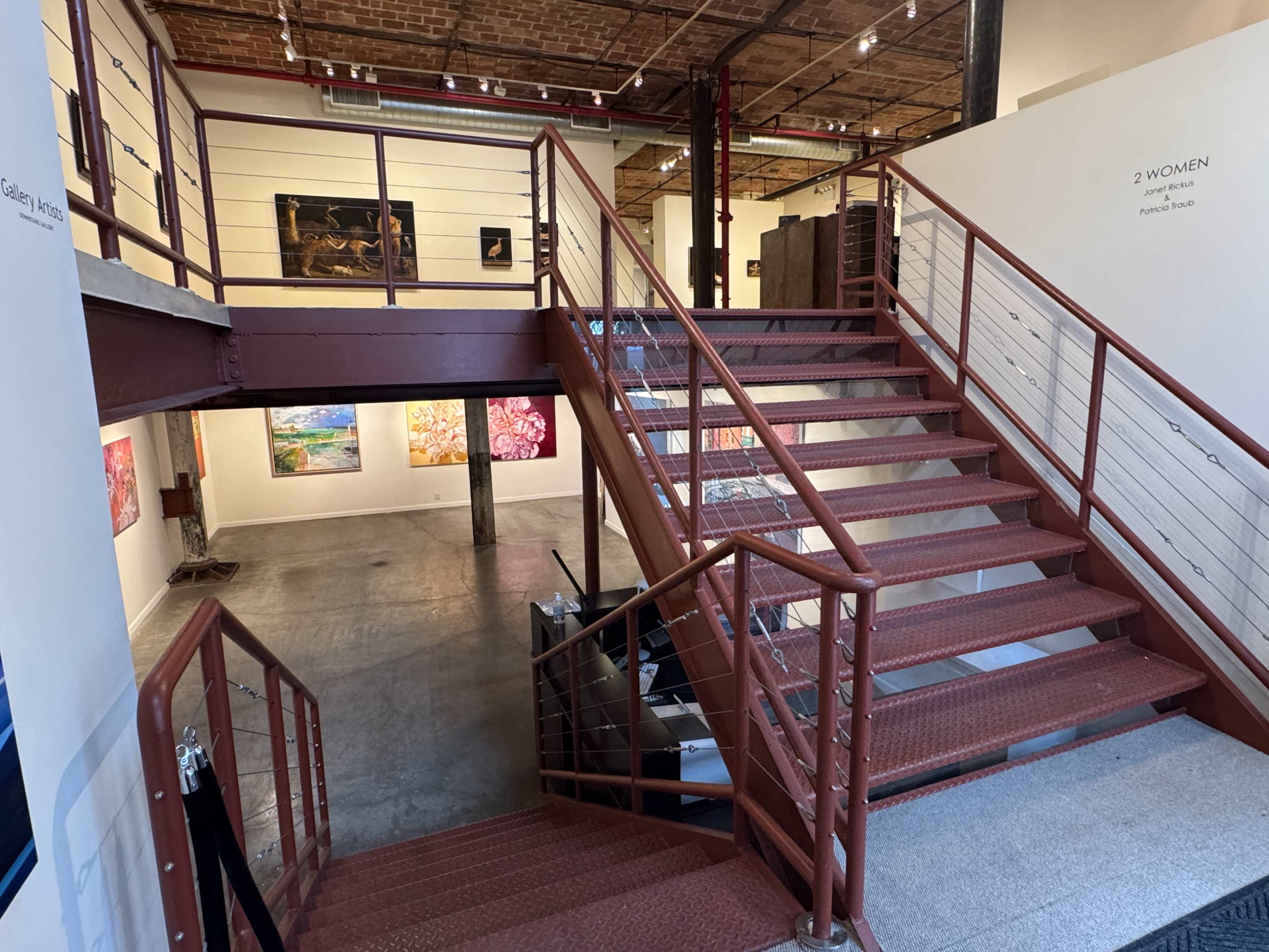 The image shows a staircase leading from the upper level to the lower level of an art gallery, with various artworks displayed on the walls.
