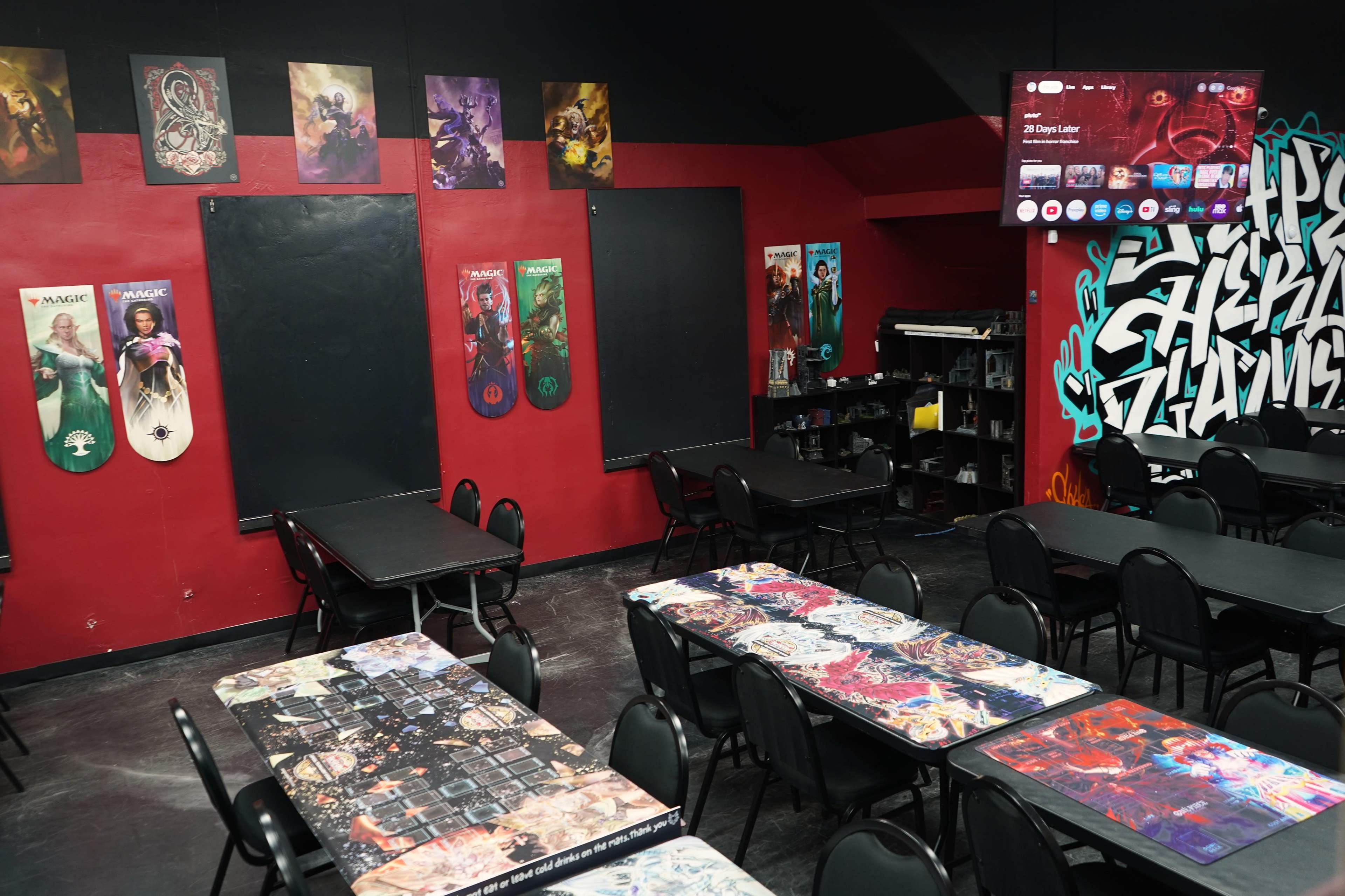 The image shows a gaming room with several tables featuring colorful designs, black chairs, and decorated walls displaying posters and a television screen.