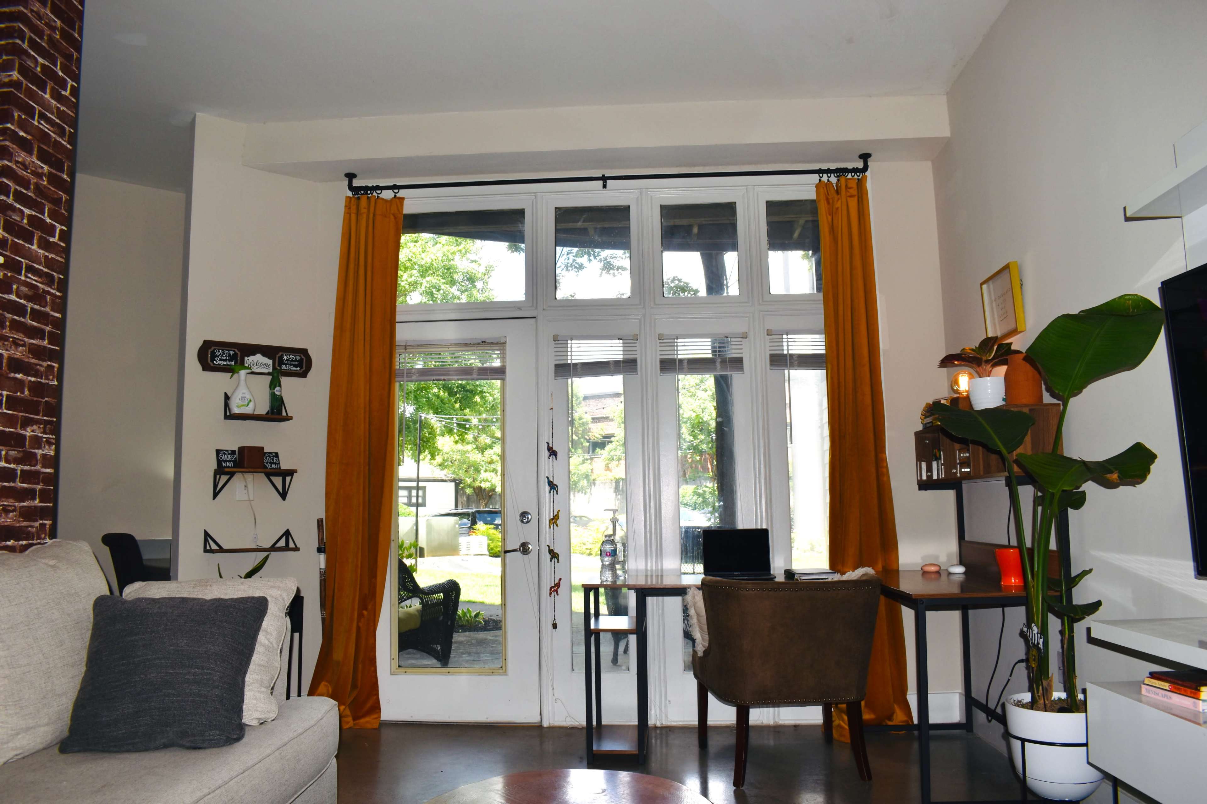 A cozy living space with a sofa, a desk by a large glass door, and yellow curtains framing the entrance to a garden area.