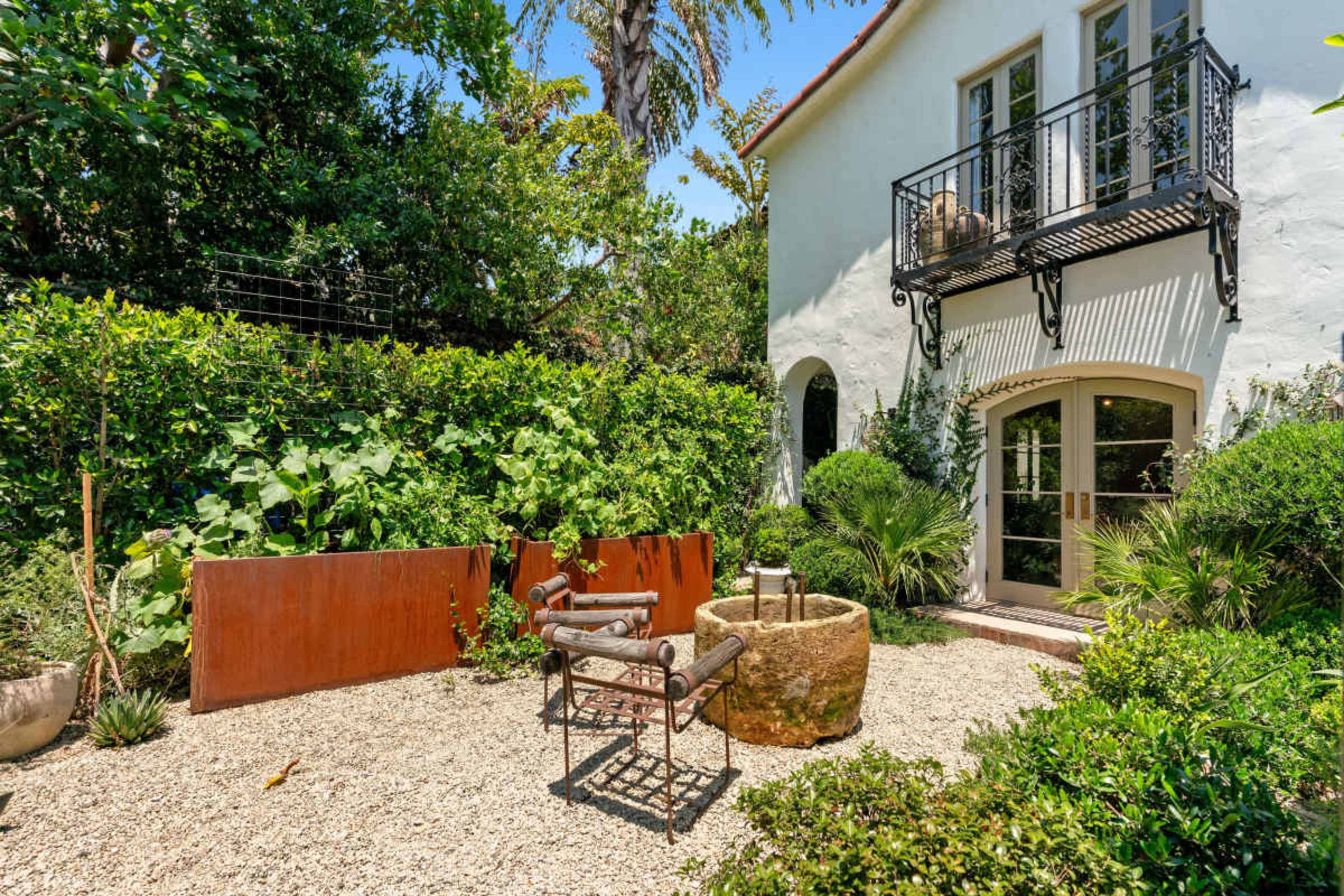 The image shows a landscaped garden area with gravel ground, lush greenery, a stone water feature, and a metal chair set near a wall.