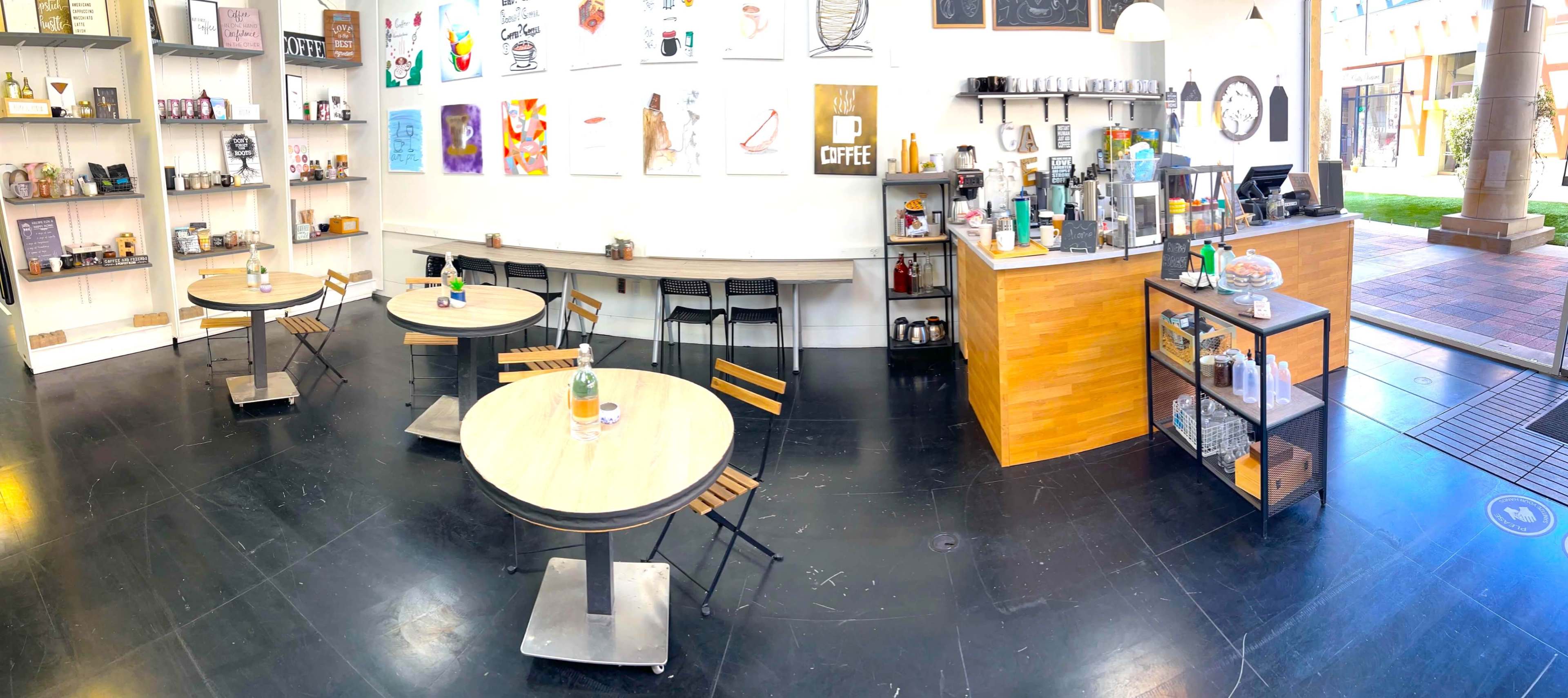 A brightly lit cafe features wooden tables with metal chairs, a service counter equipped with coffee machines, and a wall adorned with various artworks.