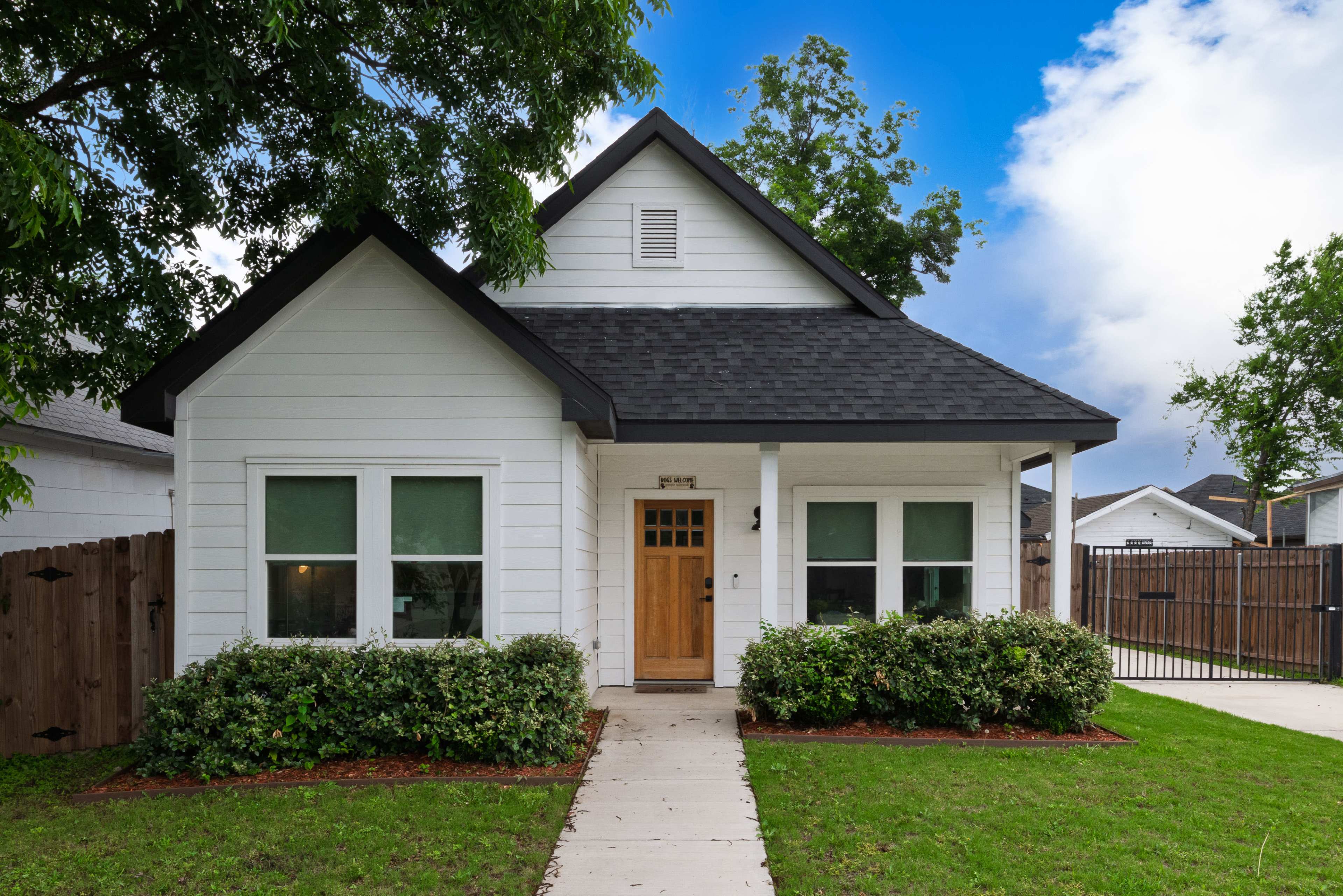 The image shows a single-story, white house with a dark roof, surrounded by neatly trimmed shrubs and a path leading to the front door.