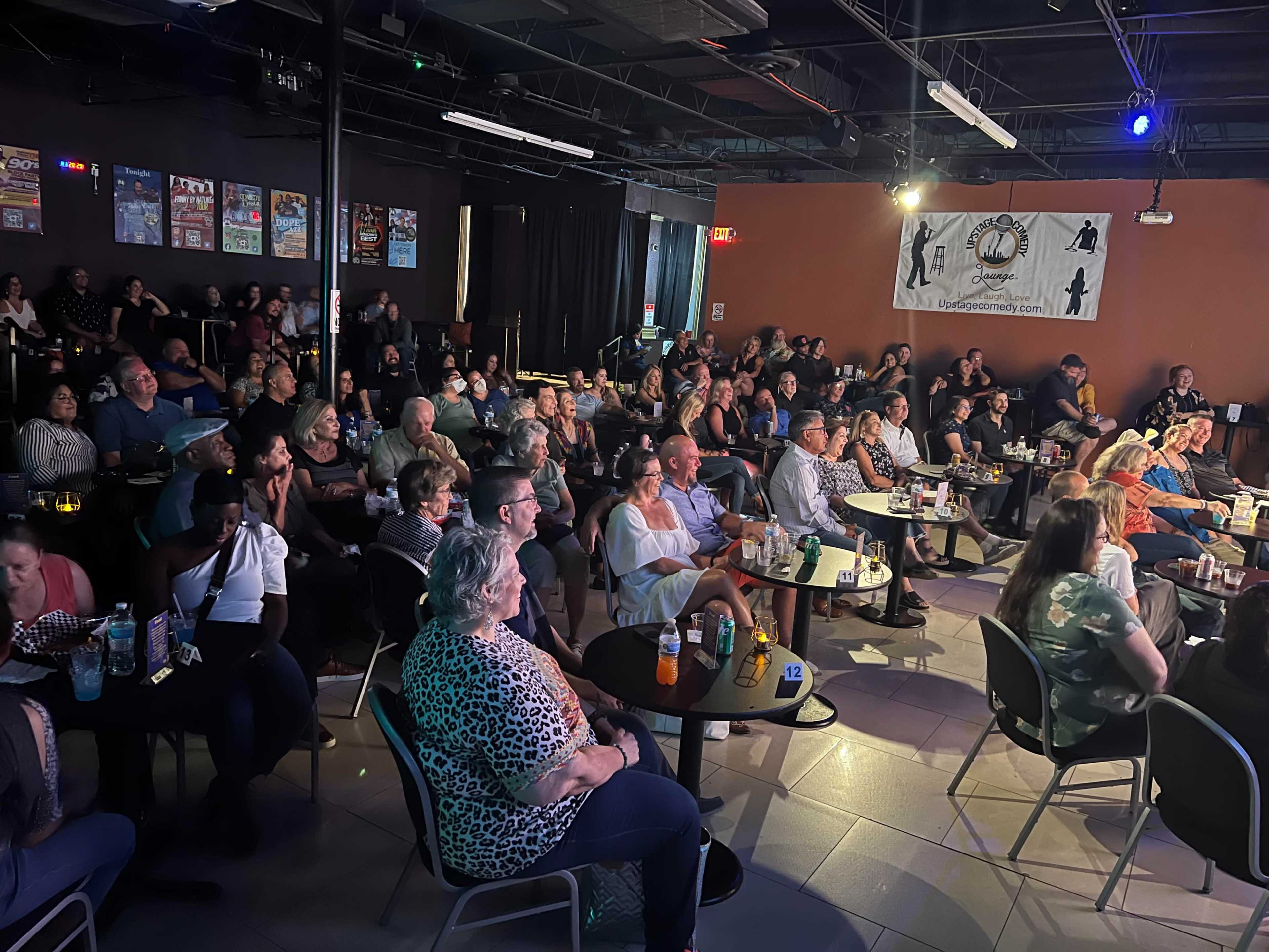 A diverse audience sits at tables, attentively watching a performance in a dimly lit venue.