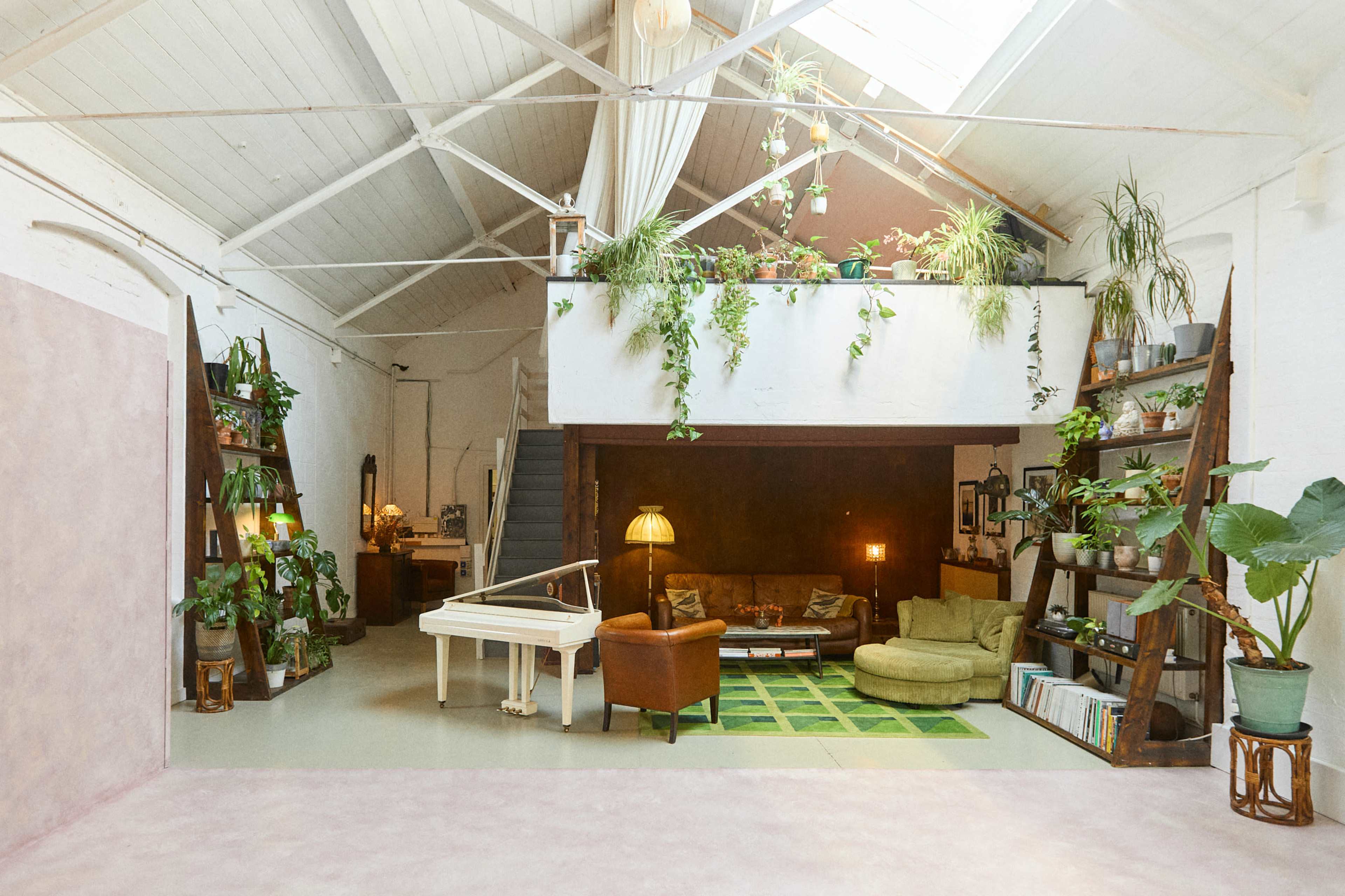 The image shows a spacious, well-lit interior of a loft-style living area featuring greenery, a piano, a cozy seating arrangement, and a lofted space above.
