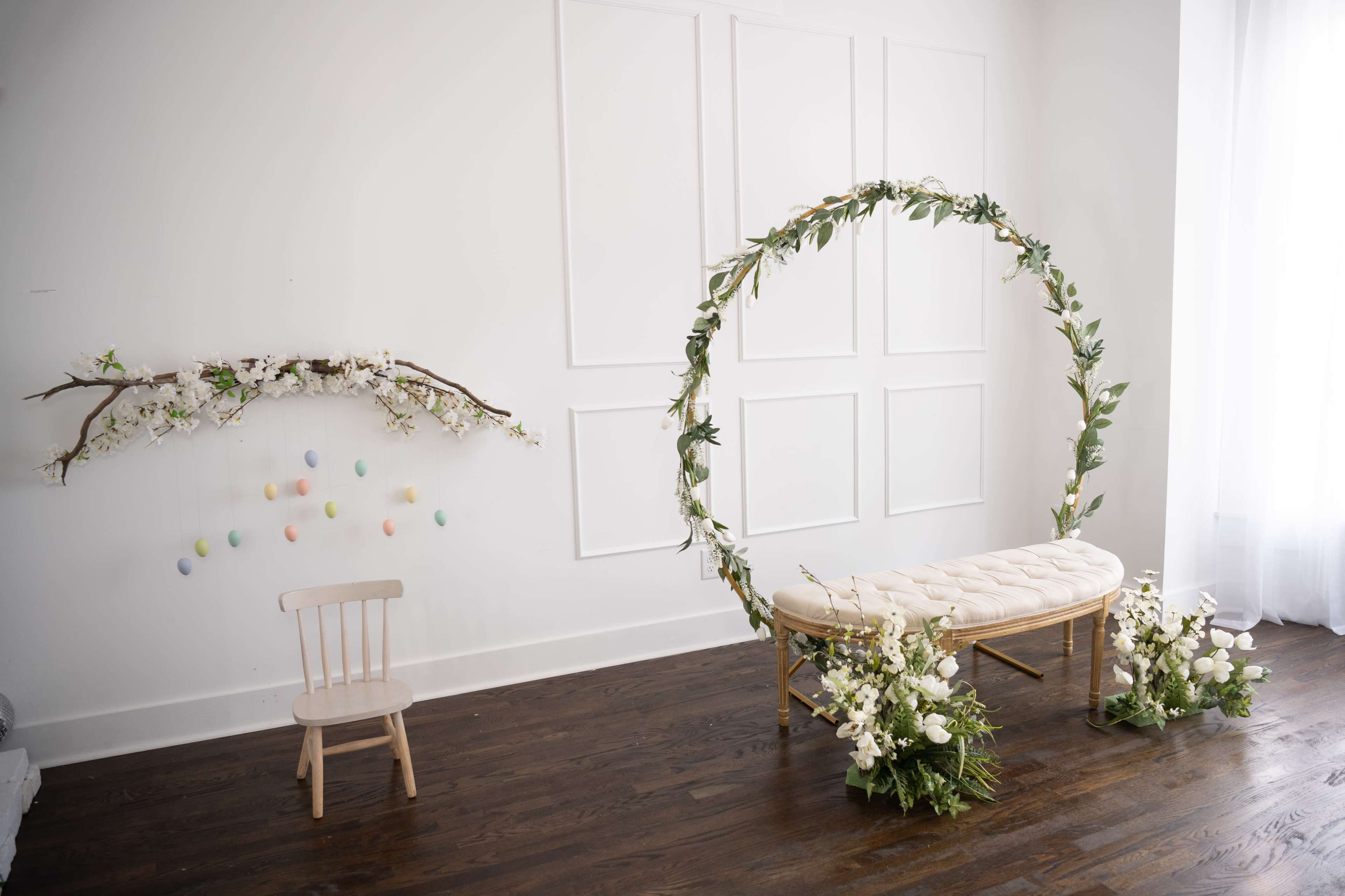 A minimalist interior features a floral arch, a tufted bench, and a wall display of decorative eggs suspended from a branch.