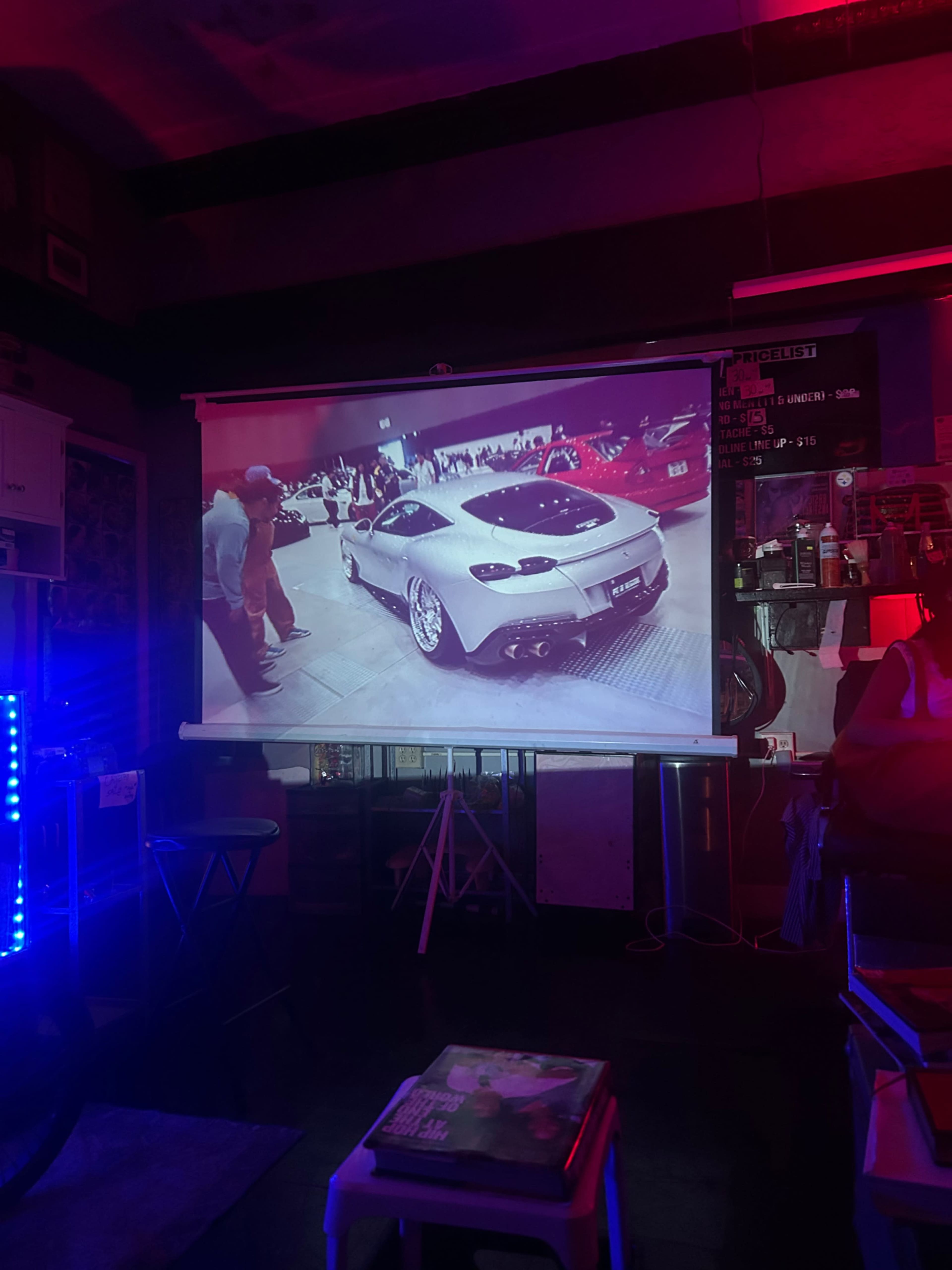 A projection screen displays an image of a modified white car with shiny wheels parked in an indoor venue surrounded by people.