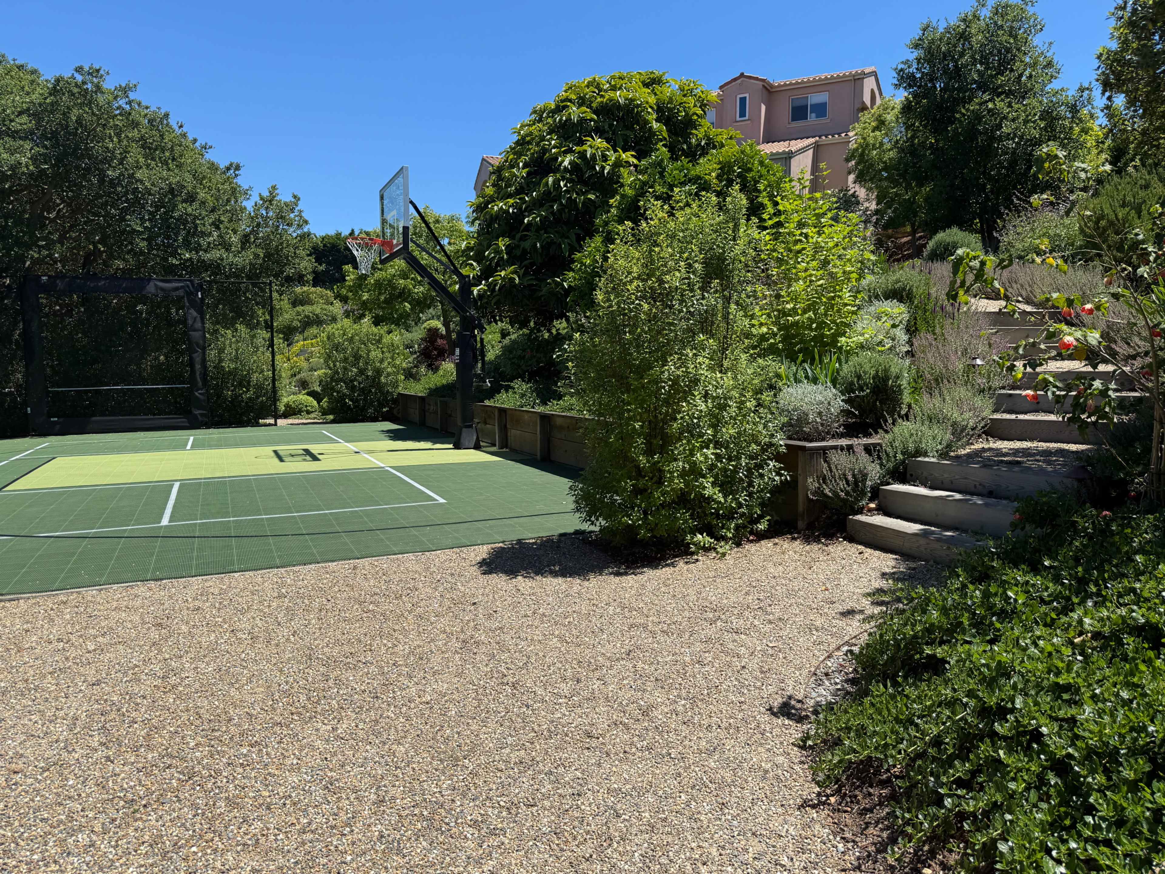 A basketball court is situated next to a landscaped garden with steps leading up to a house.