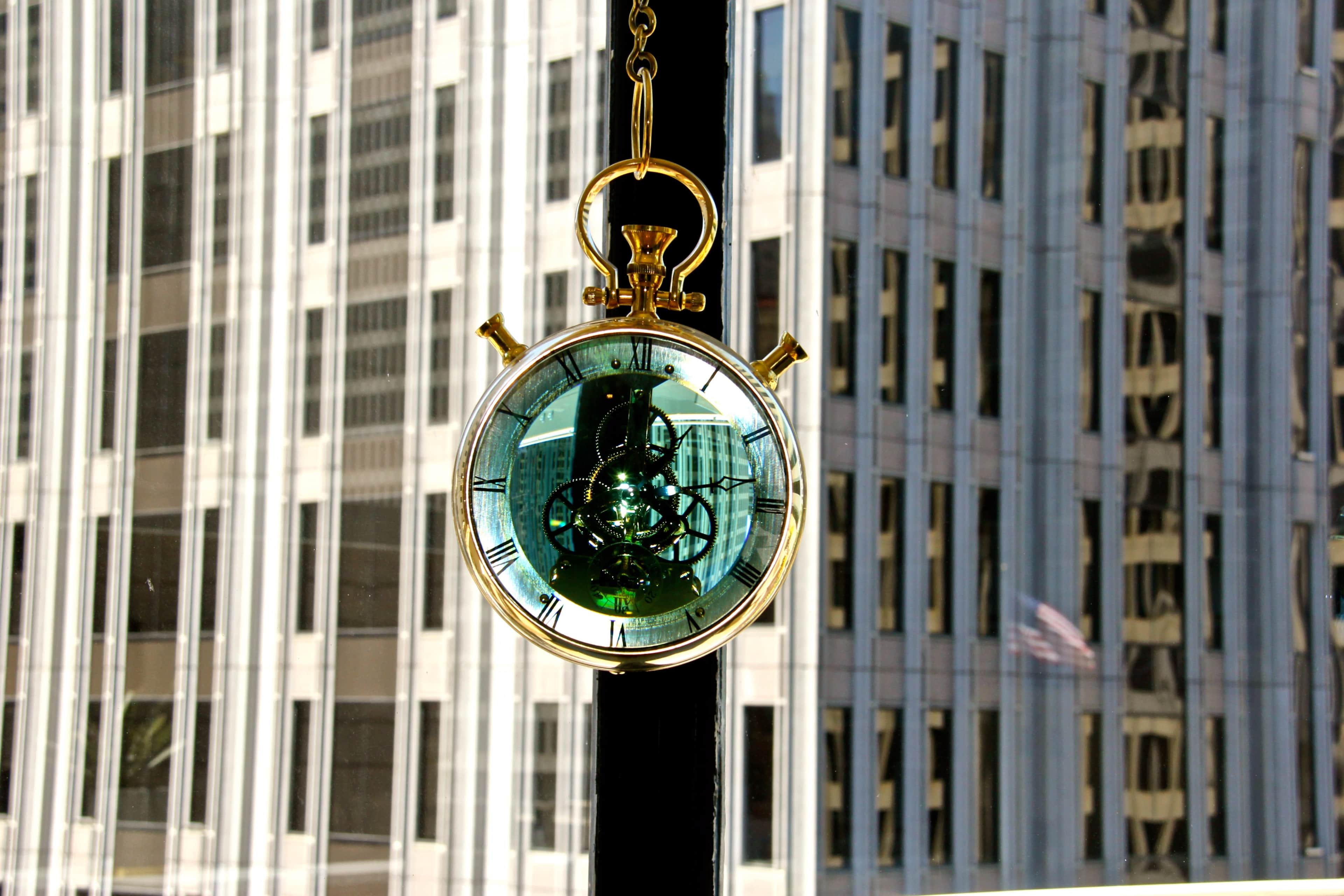 A gold pocket watch hangs in front of a window, reflecting the geometric patterns of nearby skyscrapers.