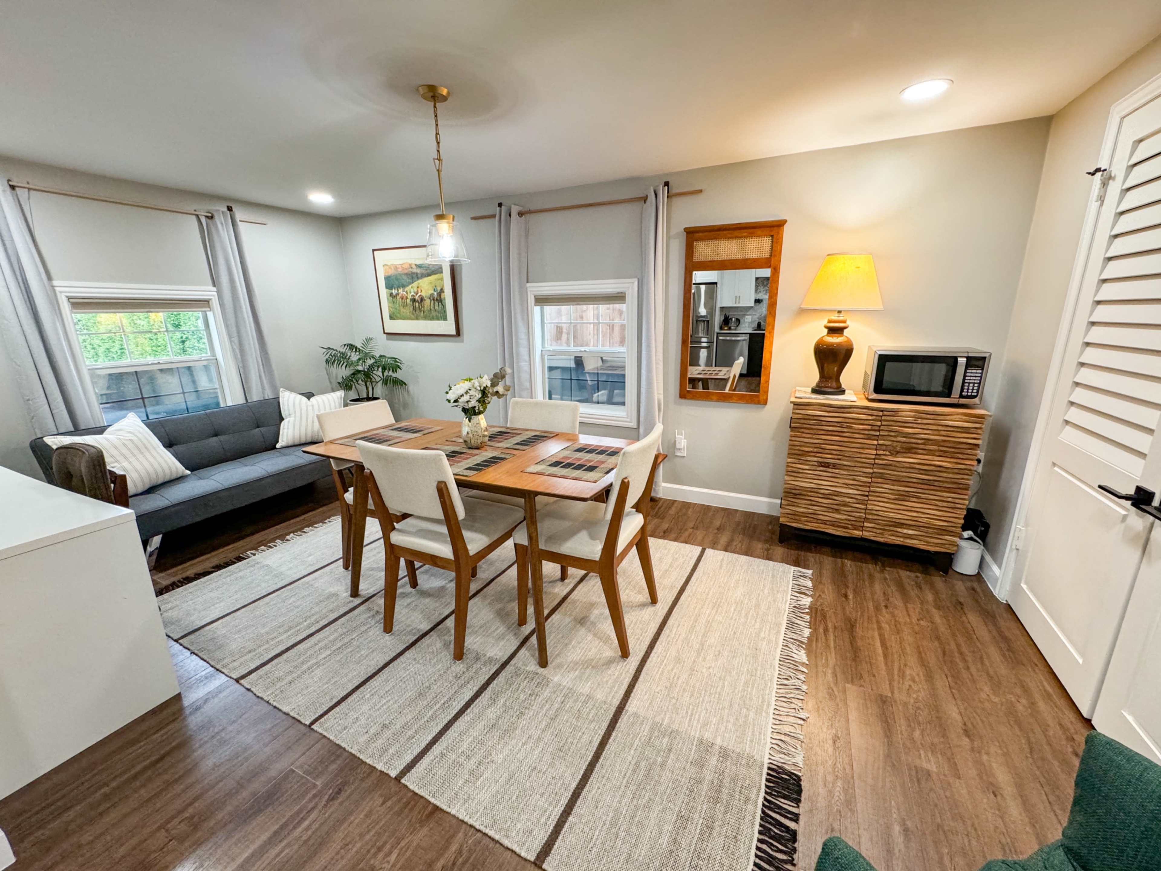A cozy living area with a sofa, dining table, and a cabinet, illuminated by a lamp and featuring large windows with natural light.