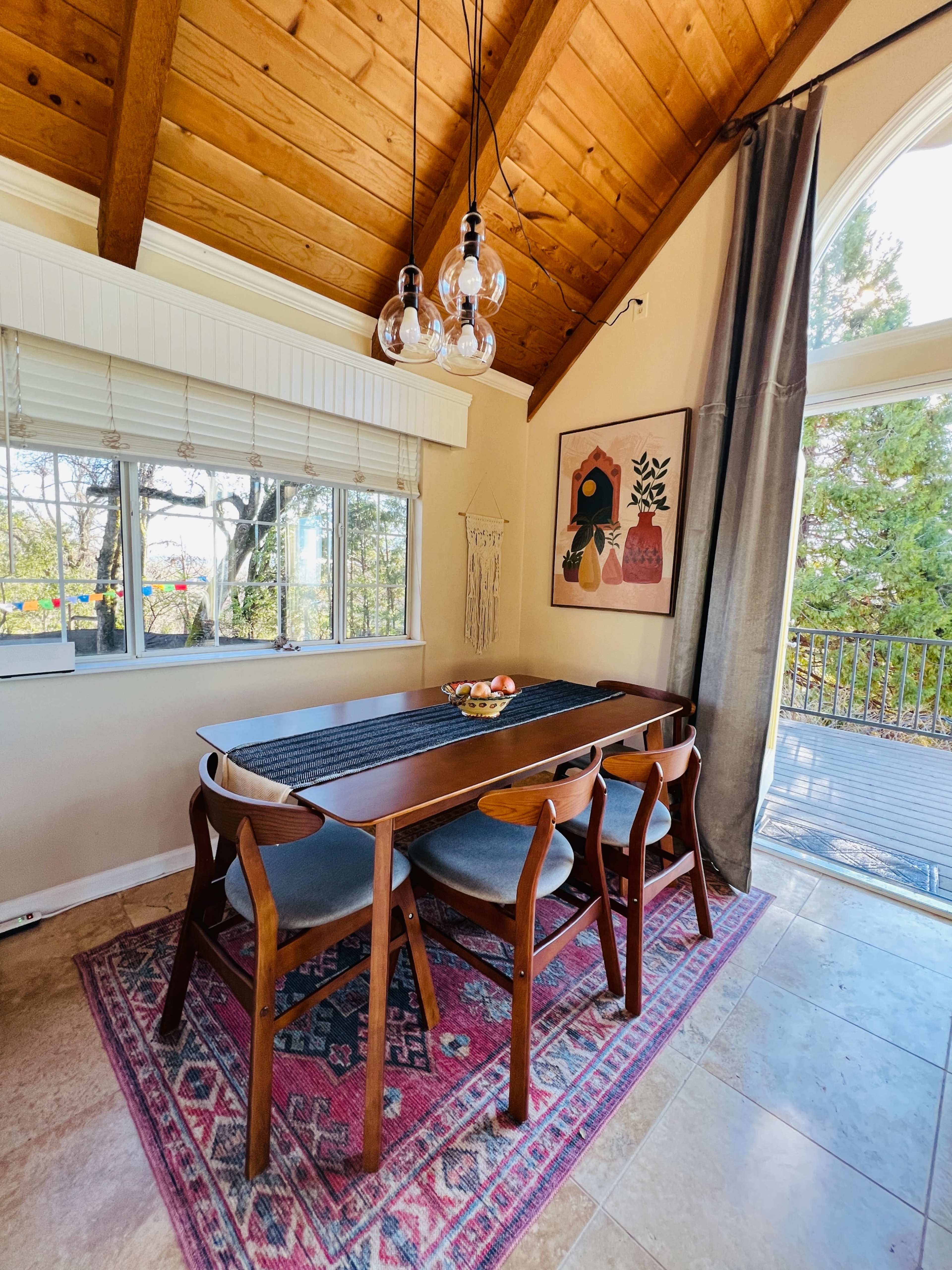 A dining area features a wooden table with four chairs, surrounded by large windows and a decorative wall hanging, under a vaulted ceiling with pendant lighting.