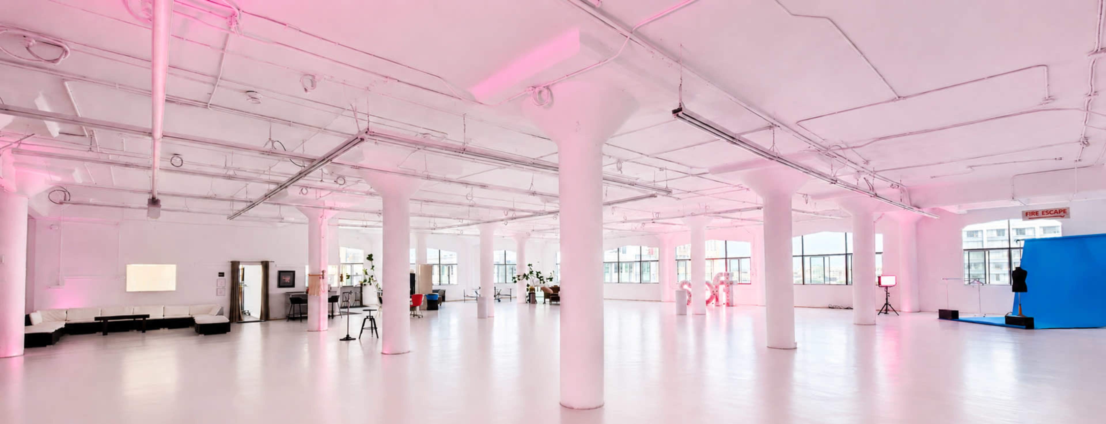 The image shows a spacious, brightly lit studio with white floors, exposed pipes, and a few decorative elements, including plants and lighting equipment.