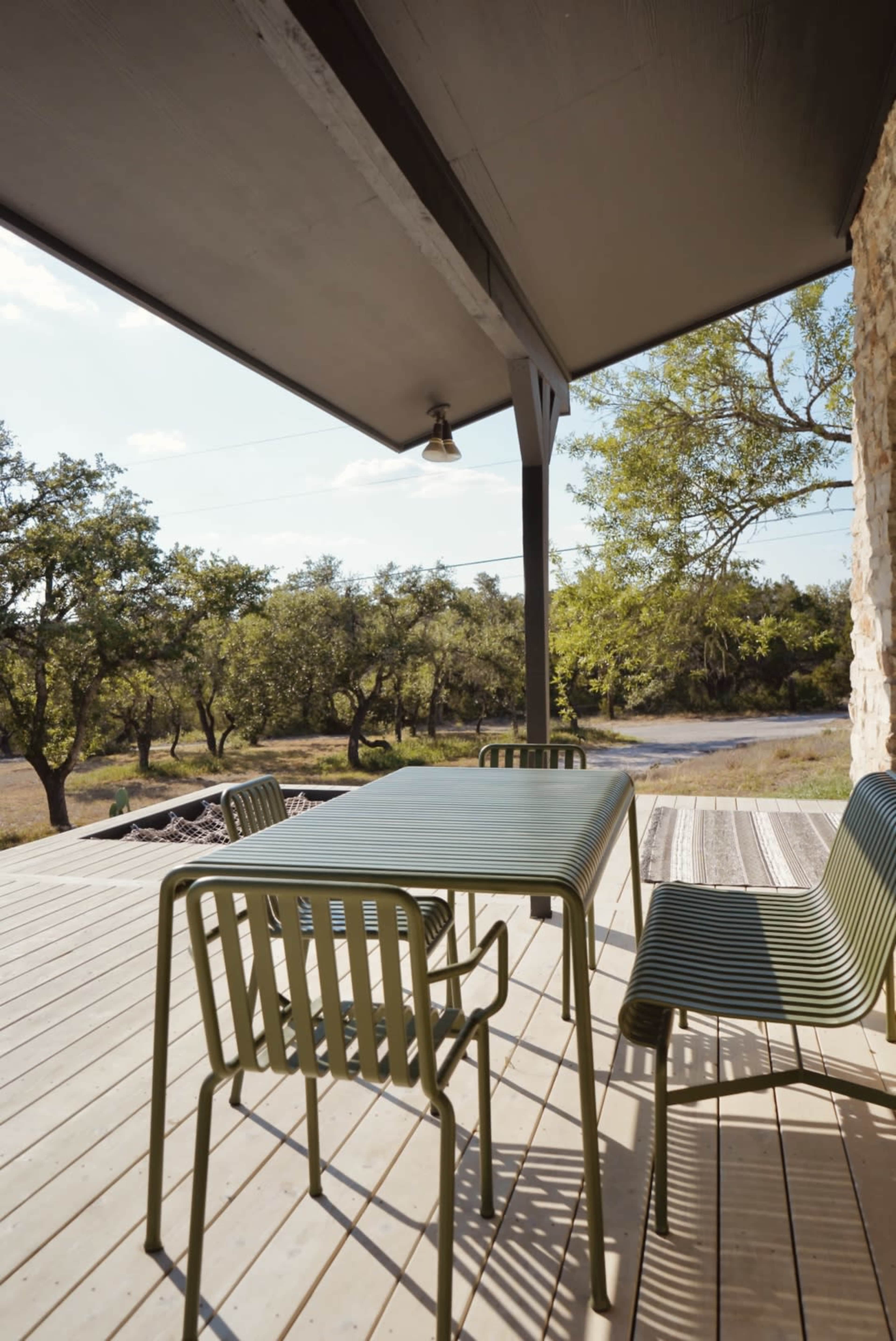 A green metal table and chairs are set on a wooden deck, surrounded by trees and a dirt road.