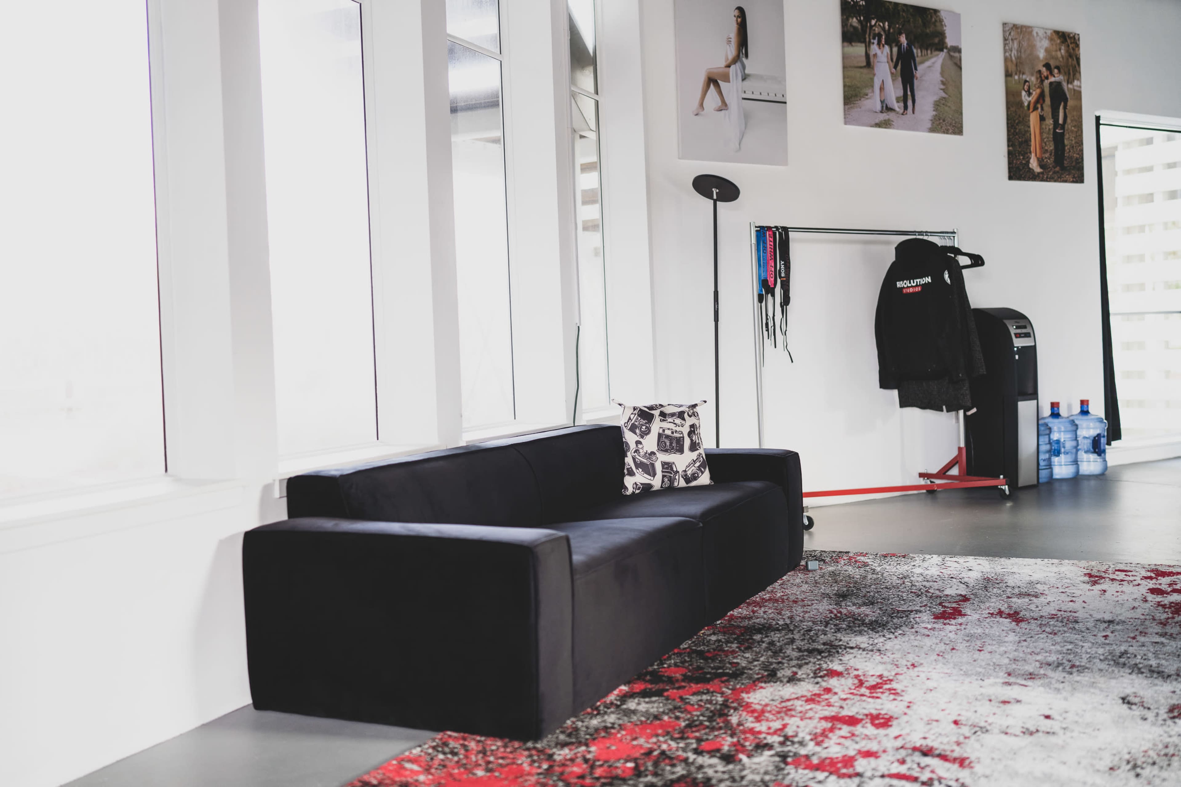 A modern, minimalist living space with a black couch, a patterned rug, and several framed photographs on the wall.