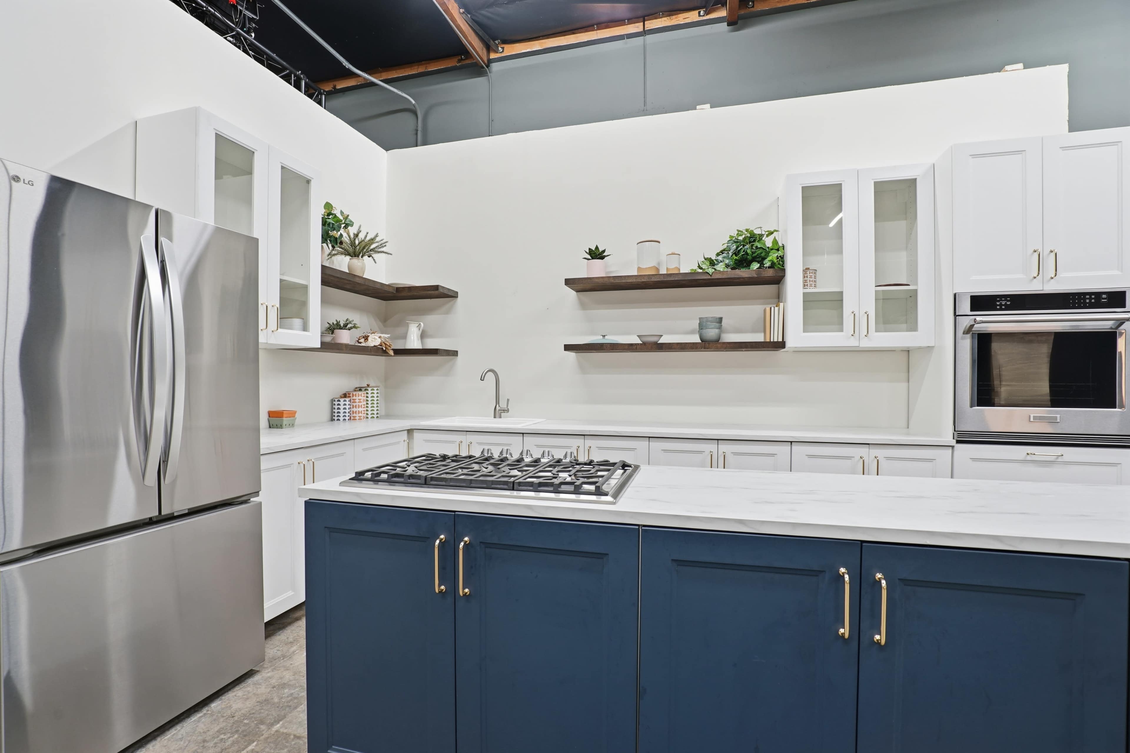 The image shows a modern kitchen featuring a stainless steel refrigerator, a gas stove on a white marble countertop, and white cabinetry with dark blue accents.