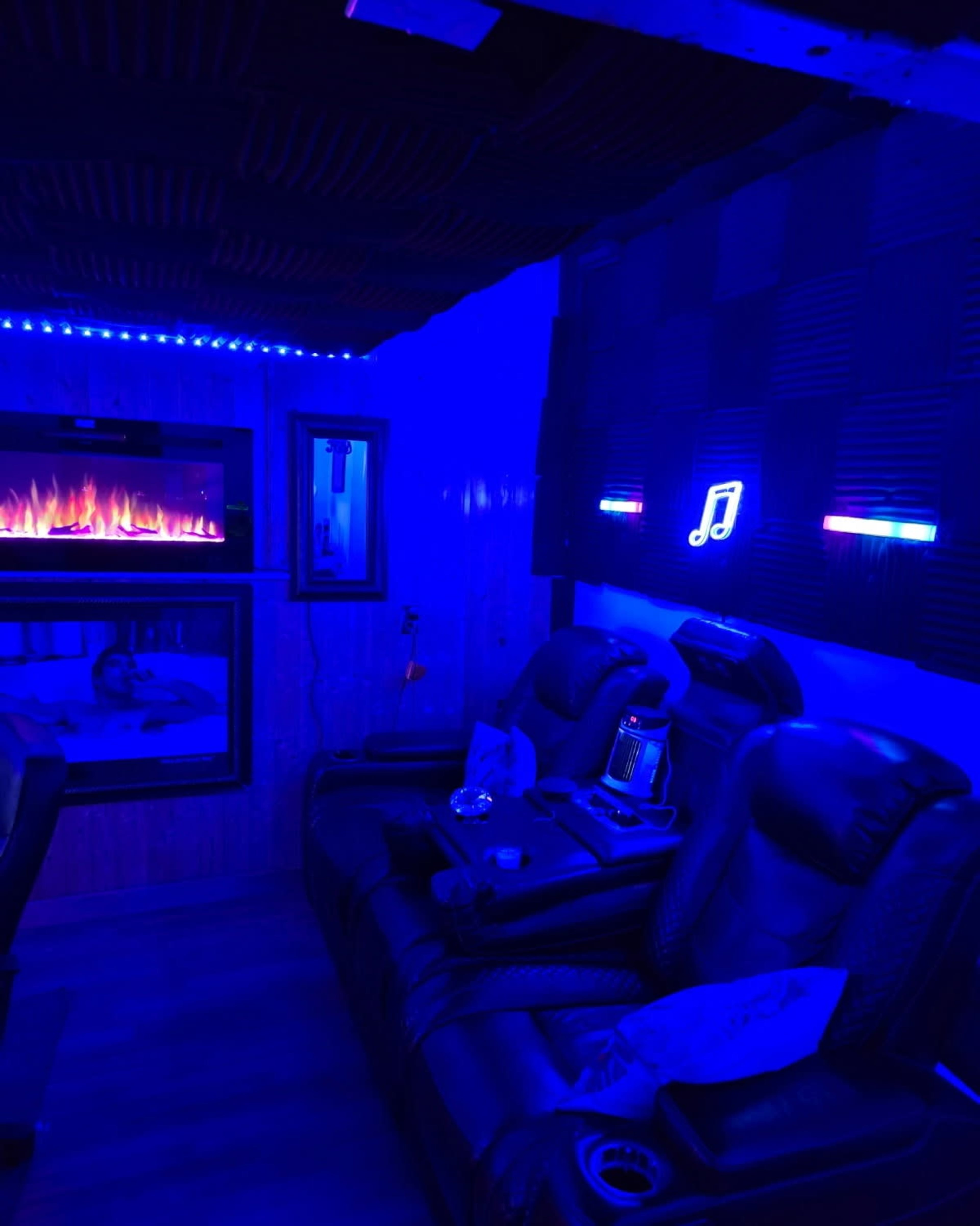 The image depicts a dimly lit room with blue lighting, featuring black leather recliners, a wall-mounted electric fireplace, and a music note decoration.