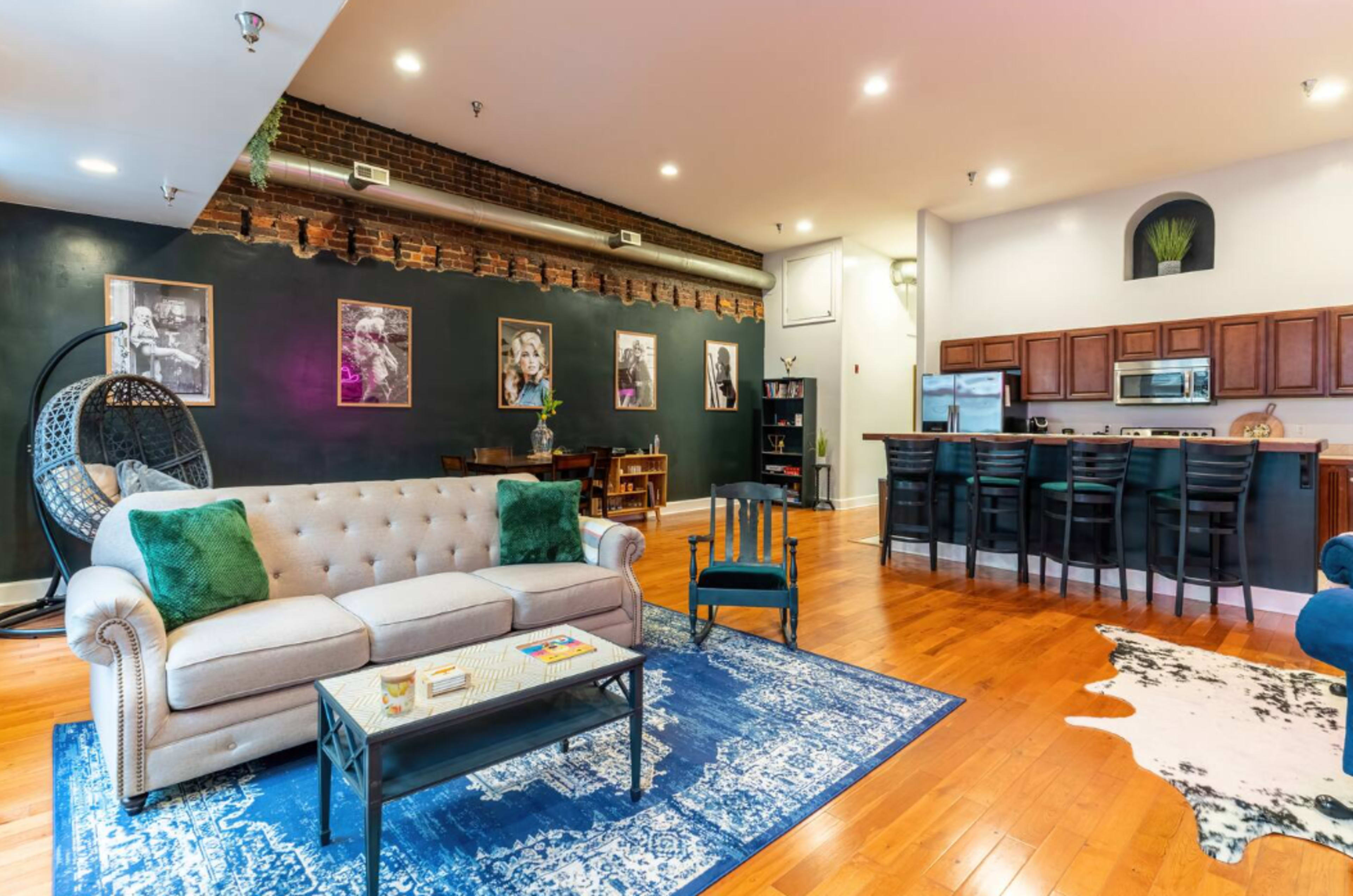 The image shows a modern living room featuring a beige tufted sofa, a blue patterned rug, and an open kitchen area with wooden cabinetry and bar seating.
