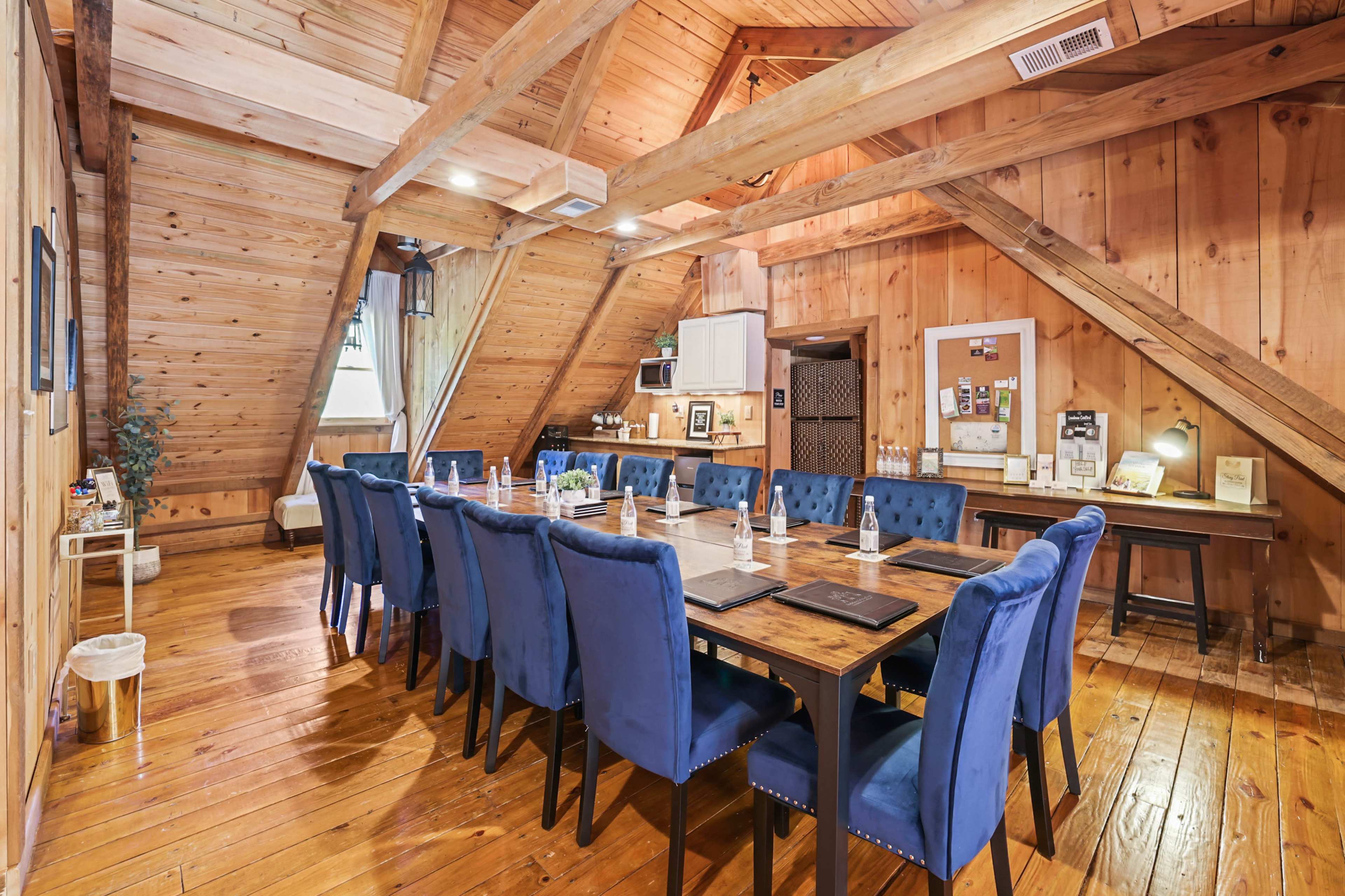 A spacious wooden meeting room features a long table surrounded by blue upholstered chairs, with exposed beams and a rustic aesthetic.