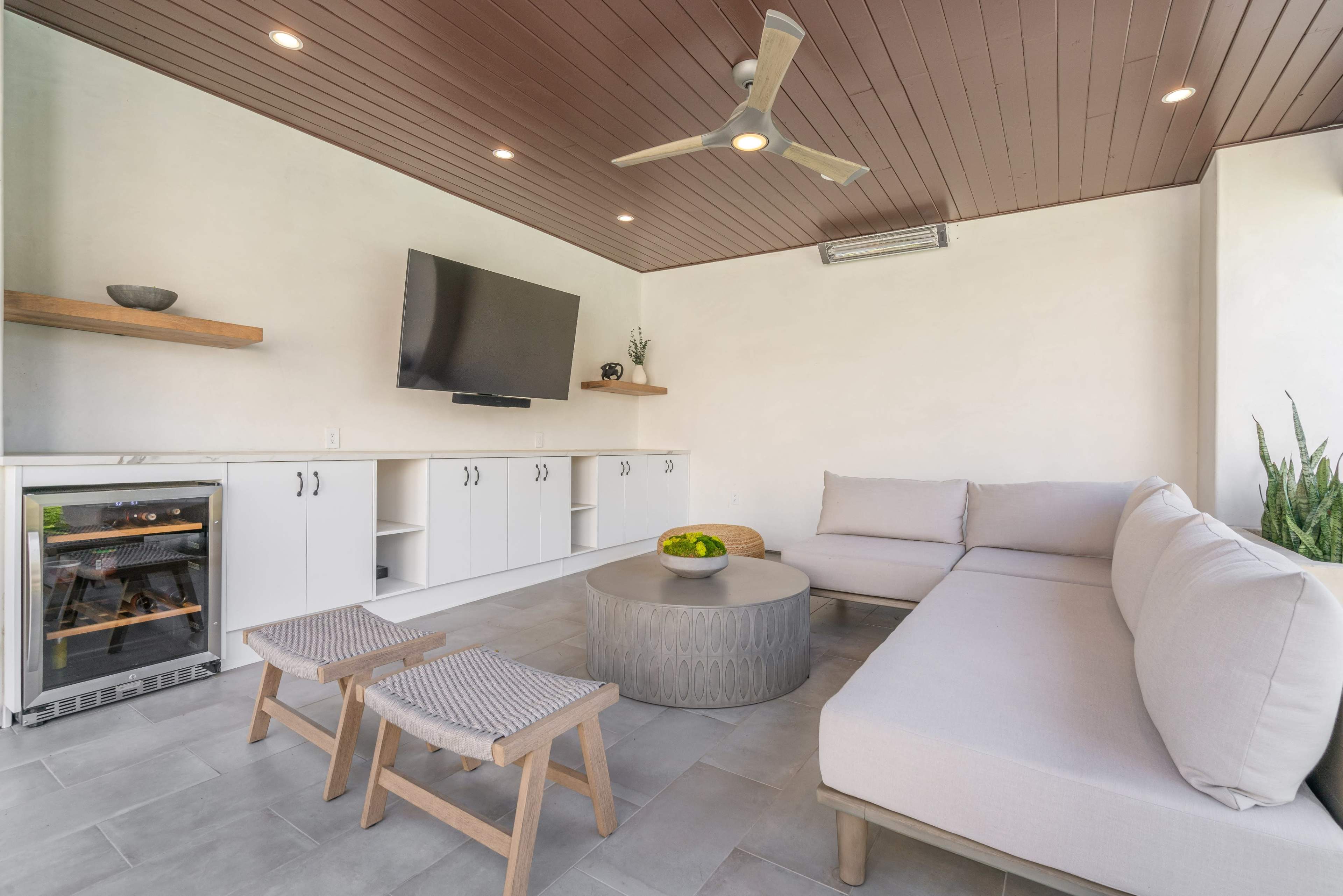 A modern indoor lounge area with a large sectional sofa, a round coffee table, two wooden stools, a wall-mounted television, and a small wine fridge.