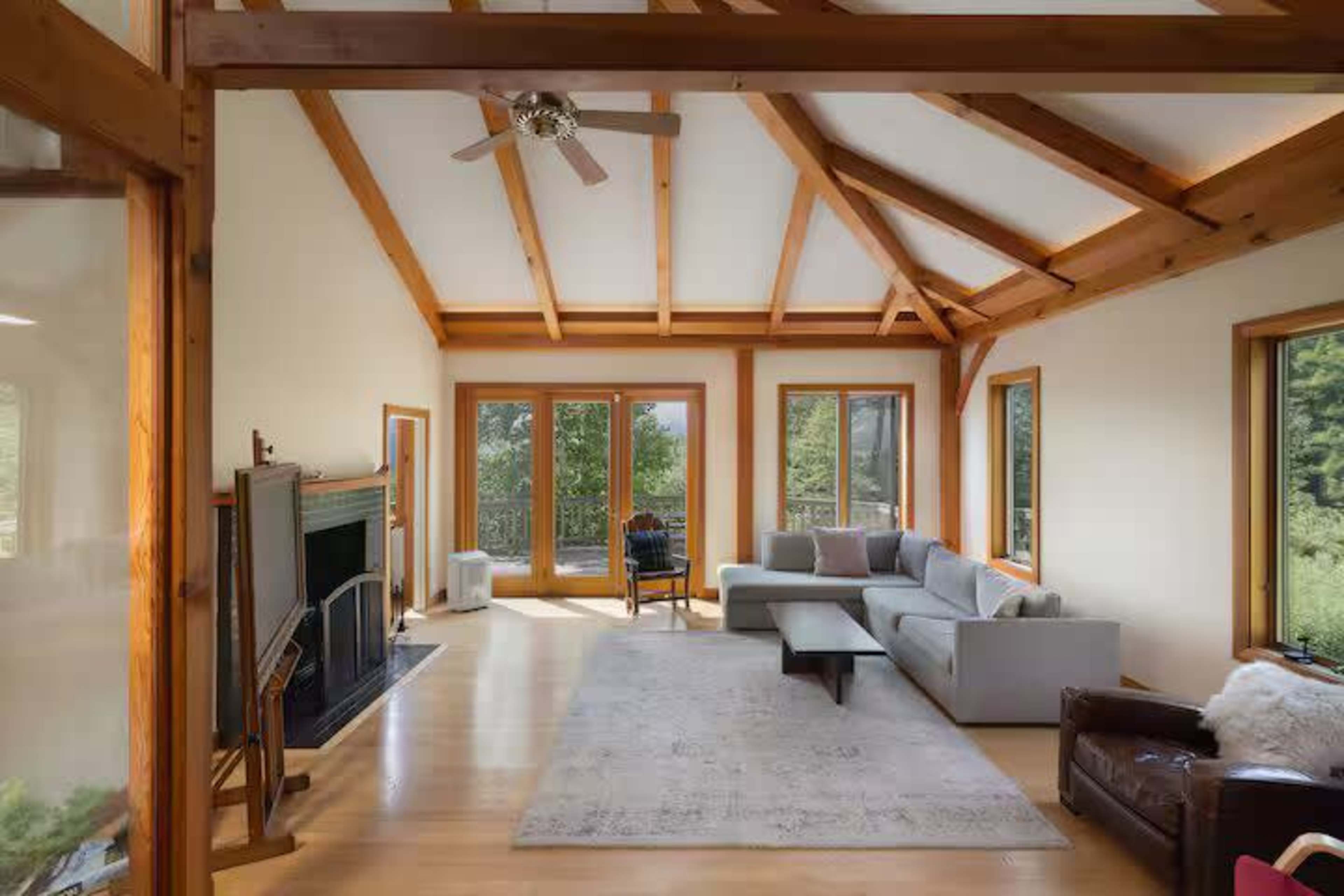 The image shows a spacious living room with a high wooden beam ceiling, large windows, a light-colored sofa, and a decorative area rug.
