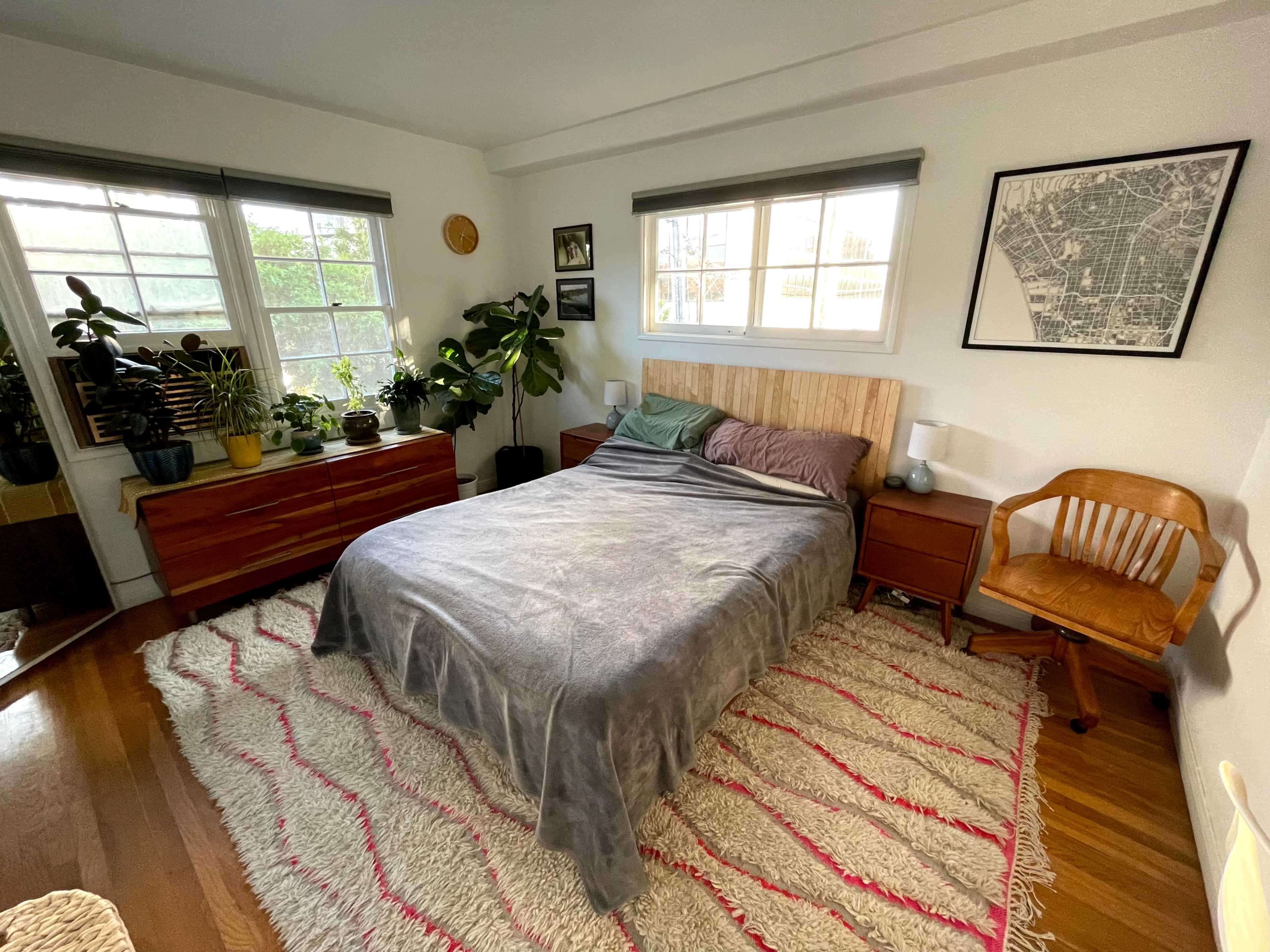 A neatly arranged bedroom with a bed, wooden nightstands, a chair, and several potted plants, all set on a textured rug.