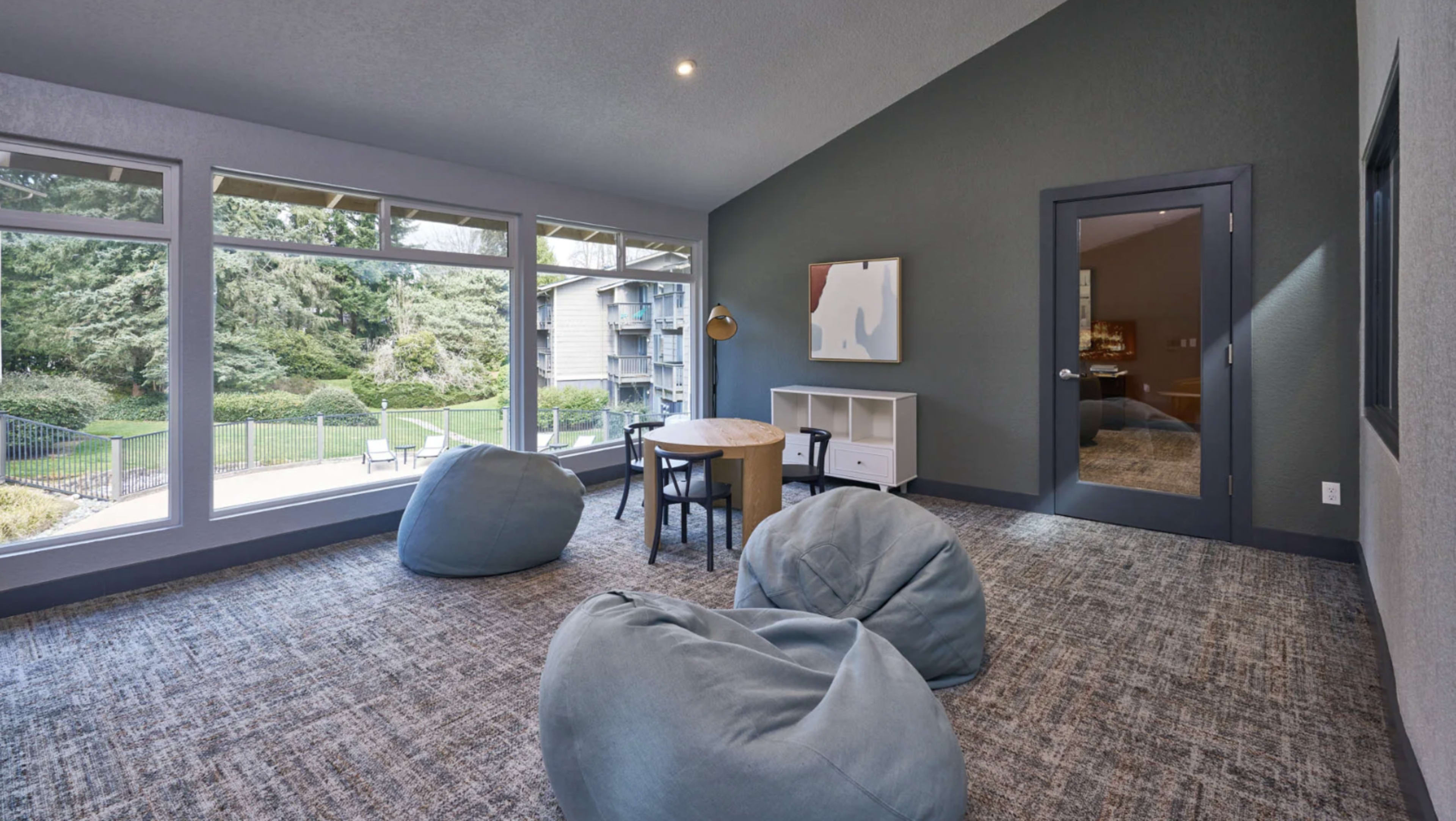 The image shows a cozy room with large windows, featuring a small round table and beanbag chairs, overlooking a green outdoor area.