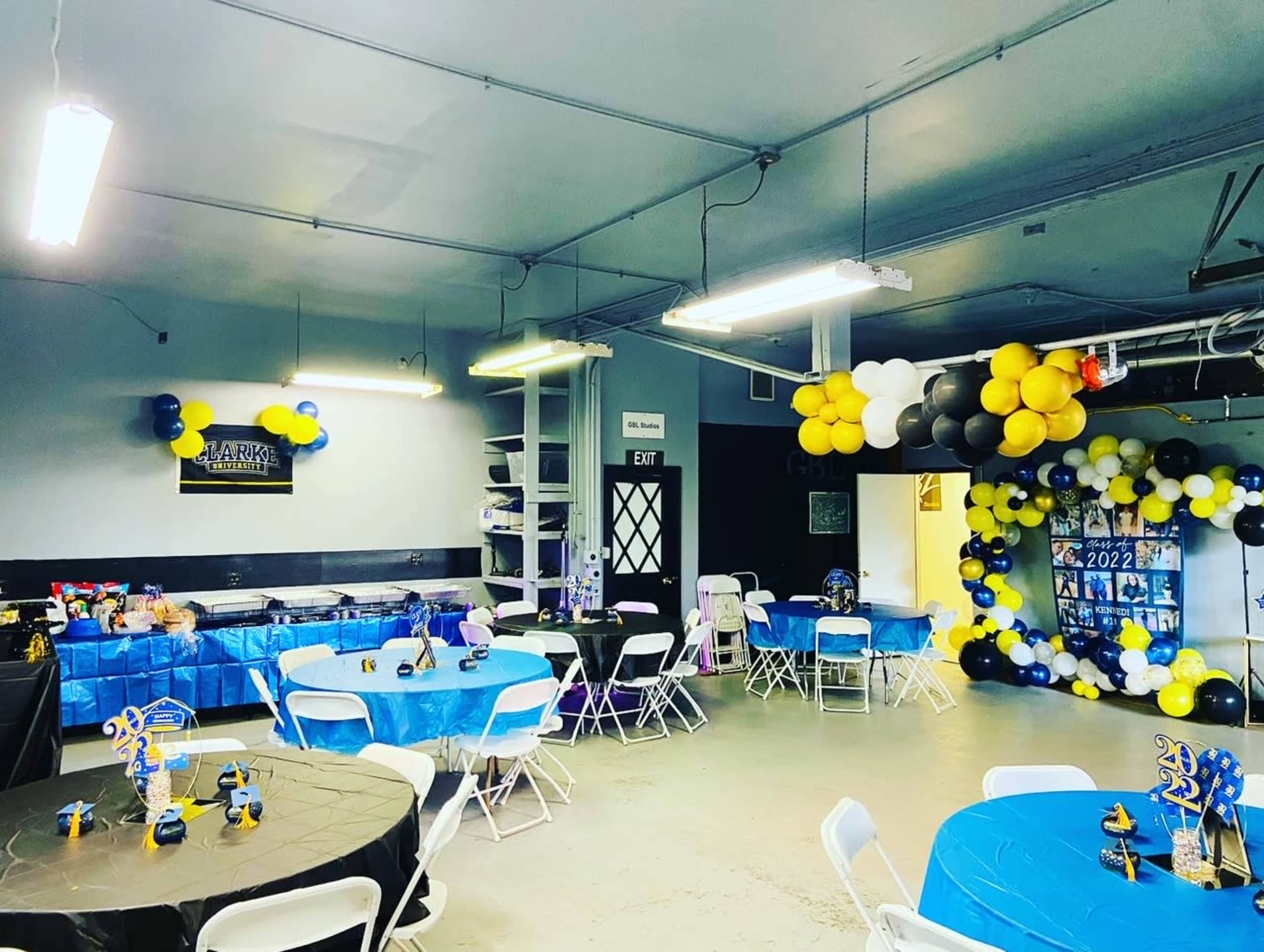 A decorated event space features blue and black tablecloths, chairs, and balloon arrangements along the walls.