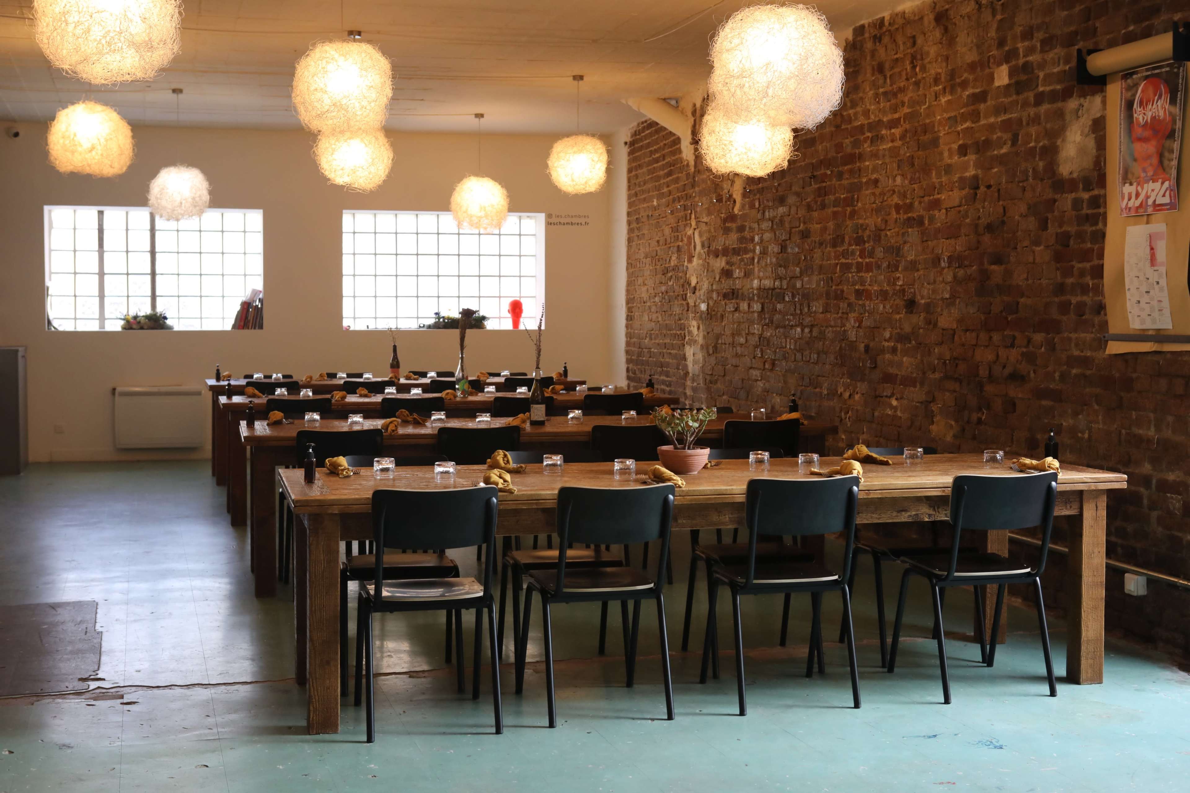 A spacious dining room features long wooden tables set with glassware and surrounded by black chairs, under illuminated pendant lights and a brick wall.