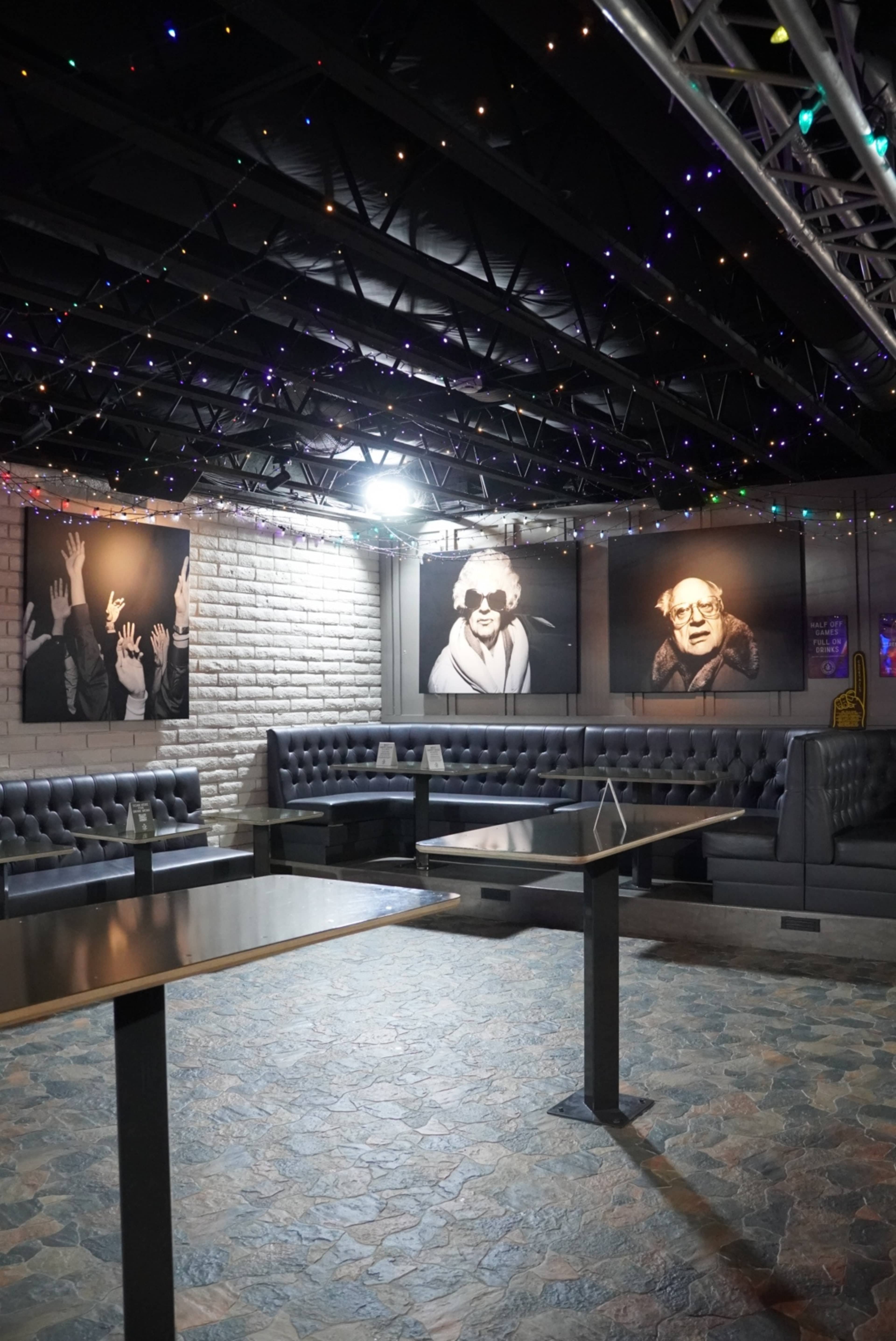The image shows a dimly lit lounge area with gray seating arrangements, illuminated by multicolored string lights and featuring large black-and-white portraits on the walls.