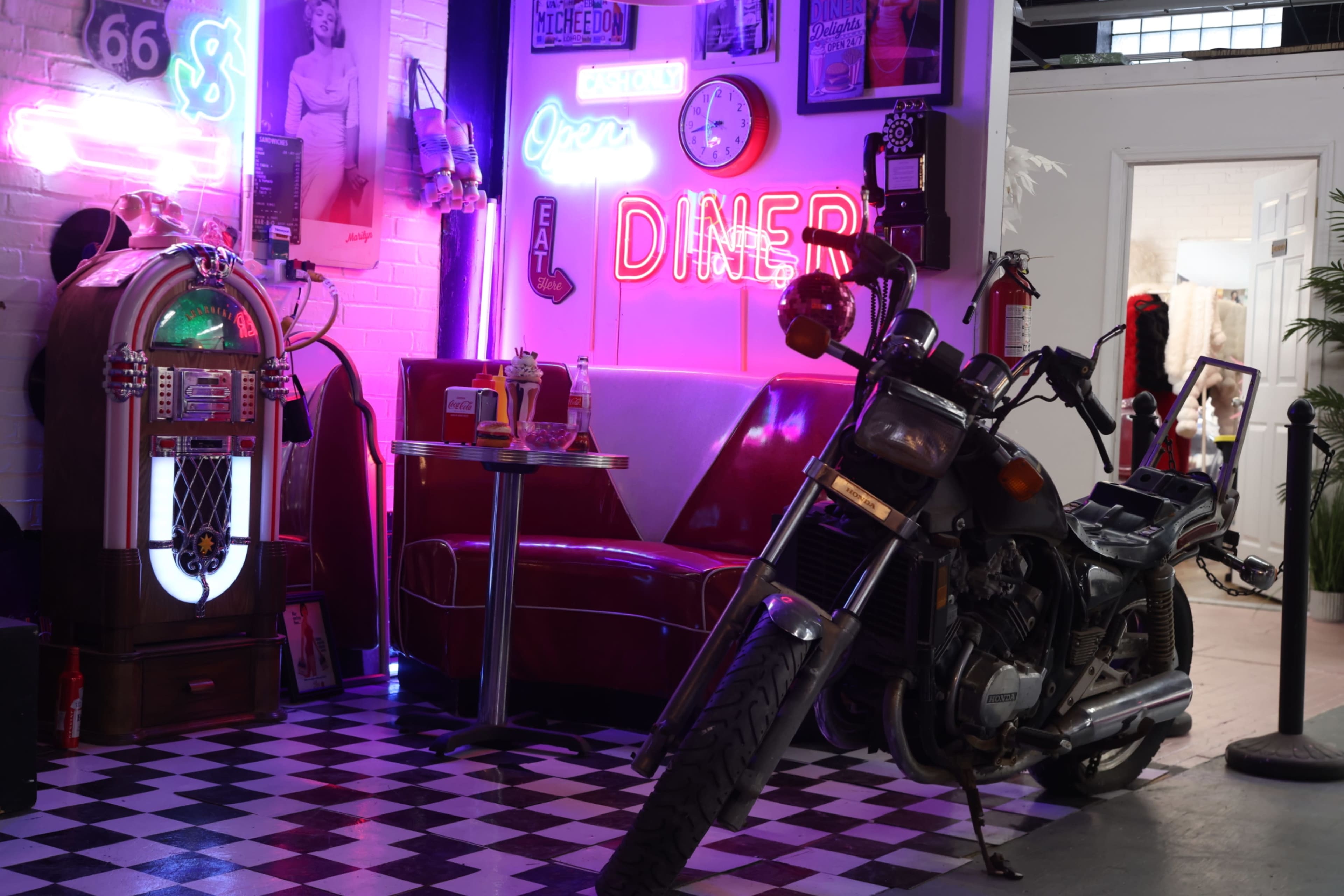 The scene features a retro diner setup with a red booth, a jukebox, neon lights, and a motorcycle positioned on a checkered floor.