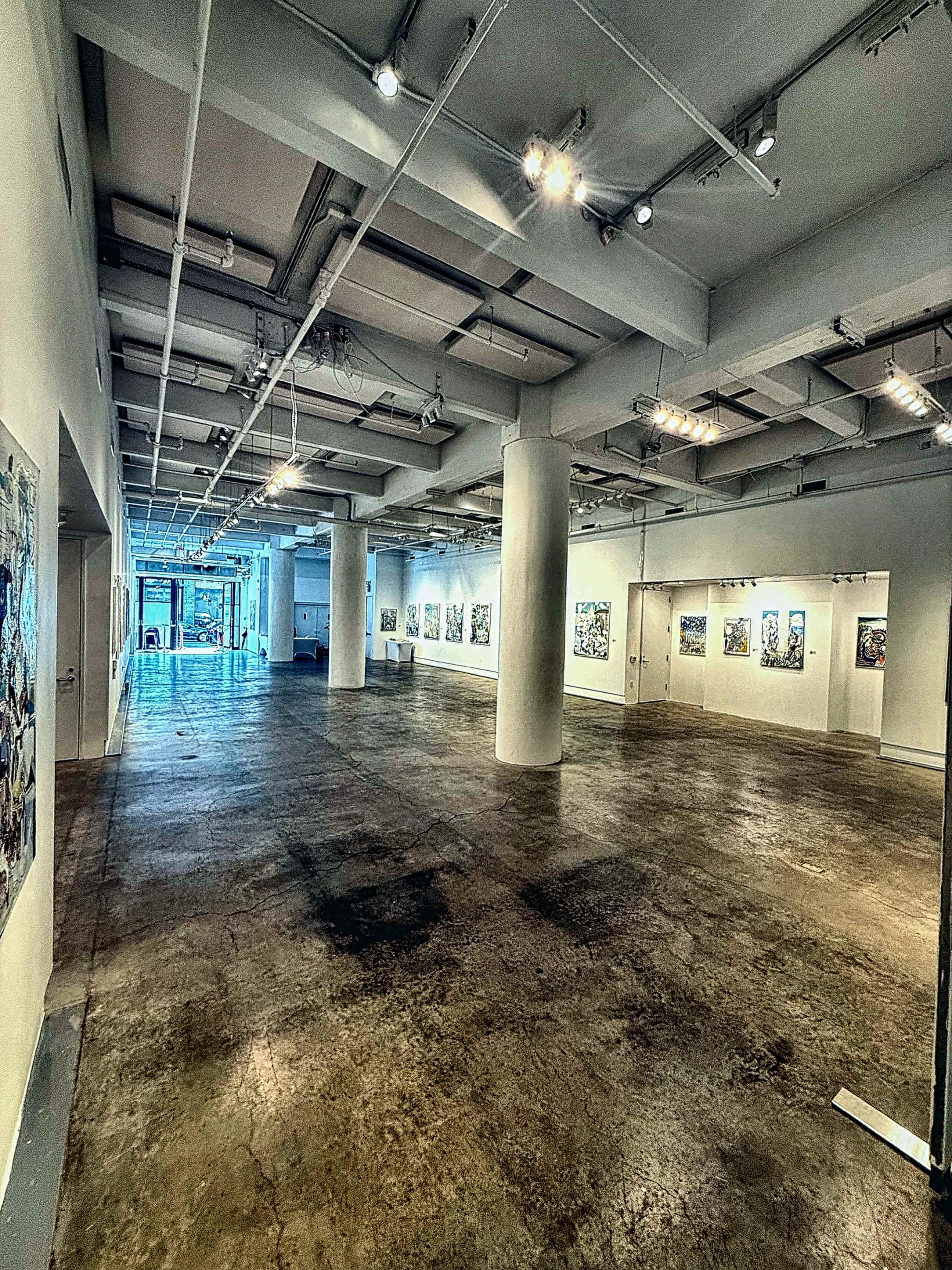 The image shows a spacious art gallery with white walls, polished concrete floors, and several artwork pieces displayed along the walls.