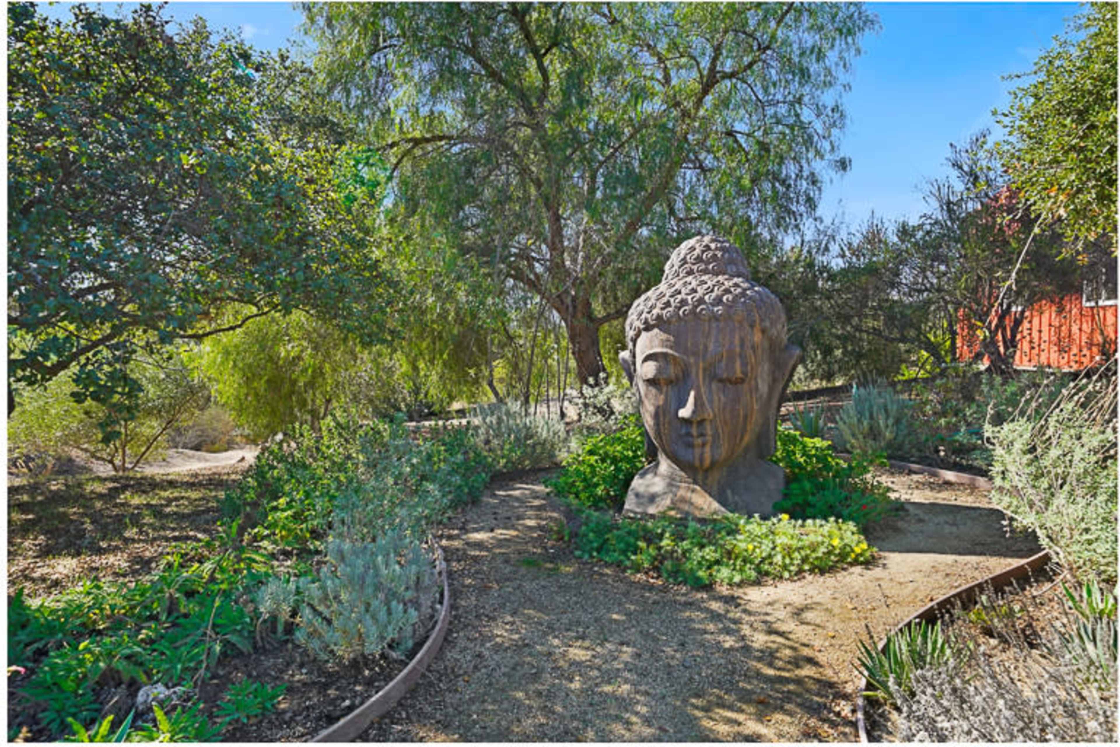 A large wooden Buddha head sculpture is surrounded by greenery in a tranquil garden setting.