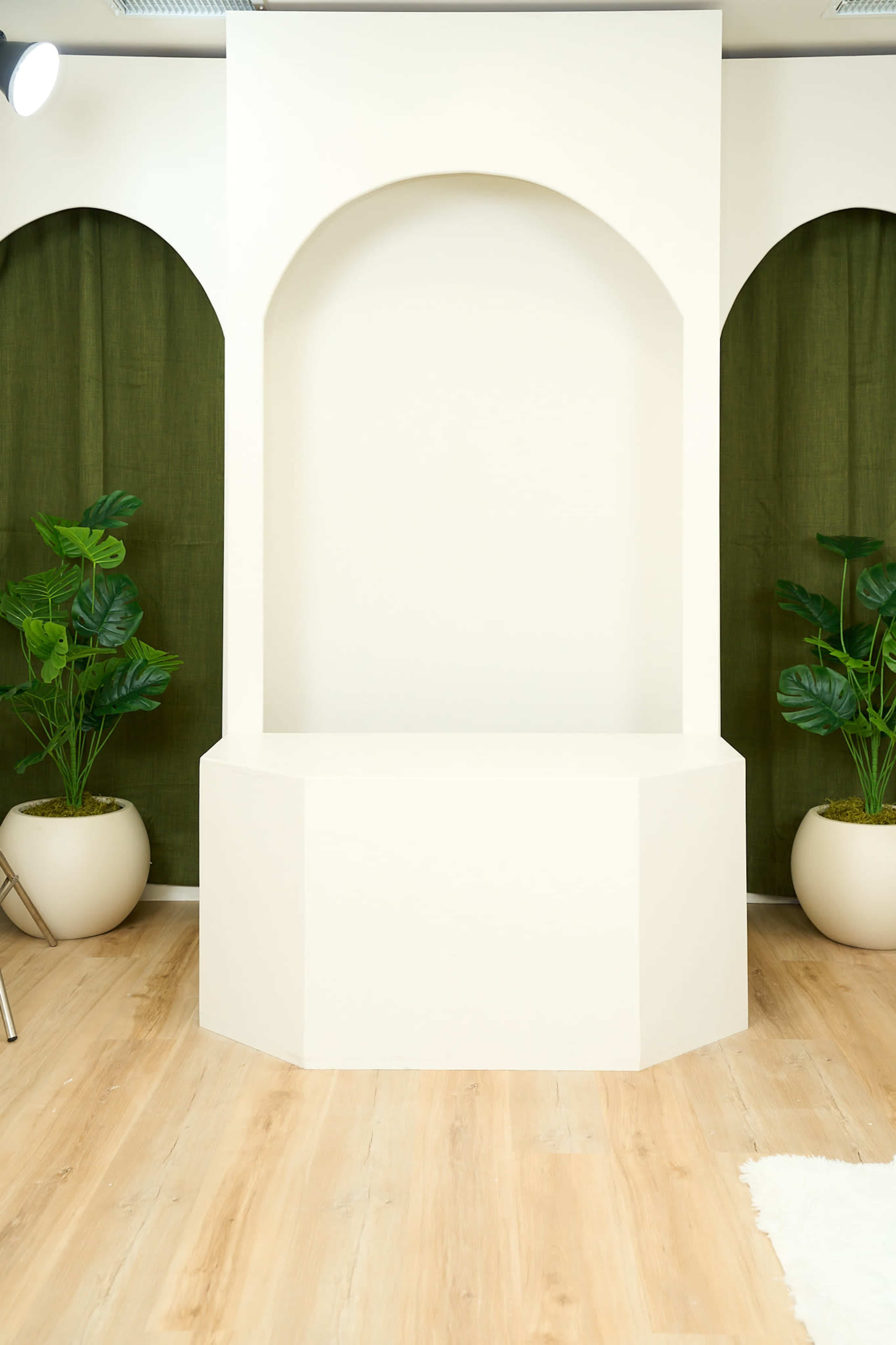 A minimalist display area with a structured, curved wall niche surrounded by green curtains and potted plants on a wooden floor.