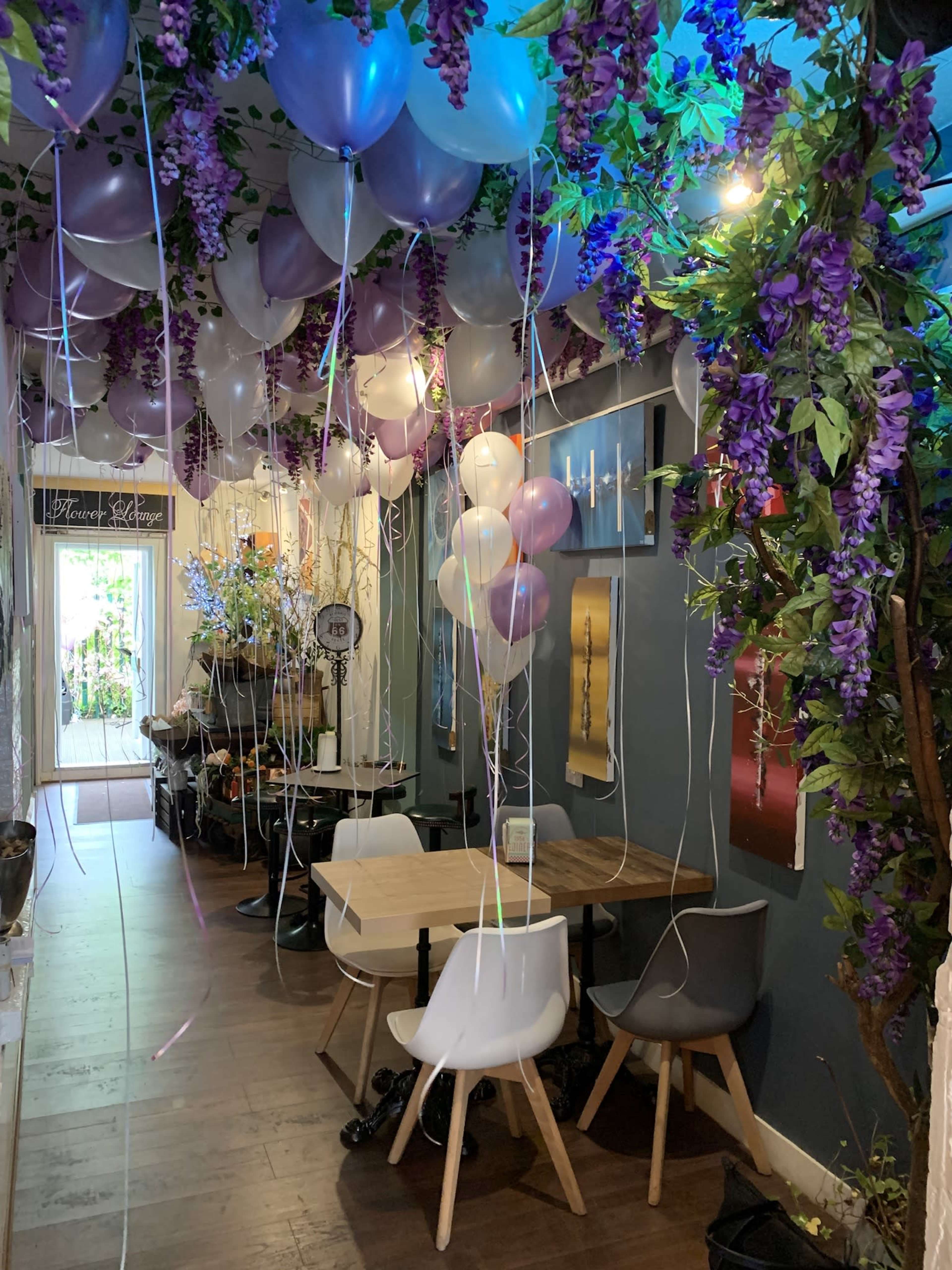 A narrow, decorated hallway features balloons and purple flowers hanging from the ceiling, with a table and chairs set up for visitors.