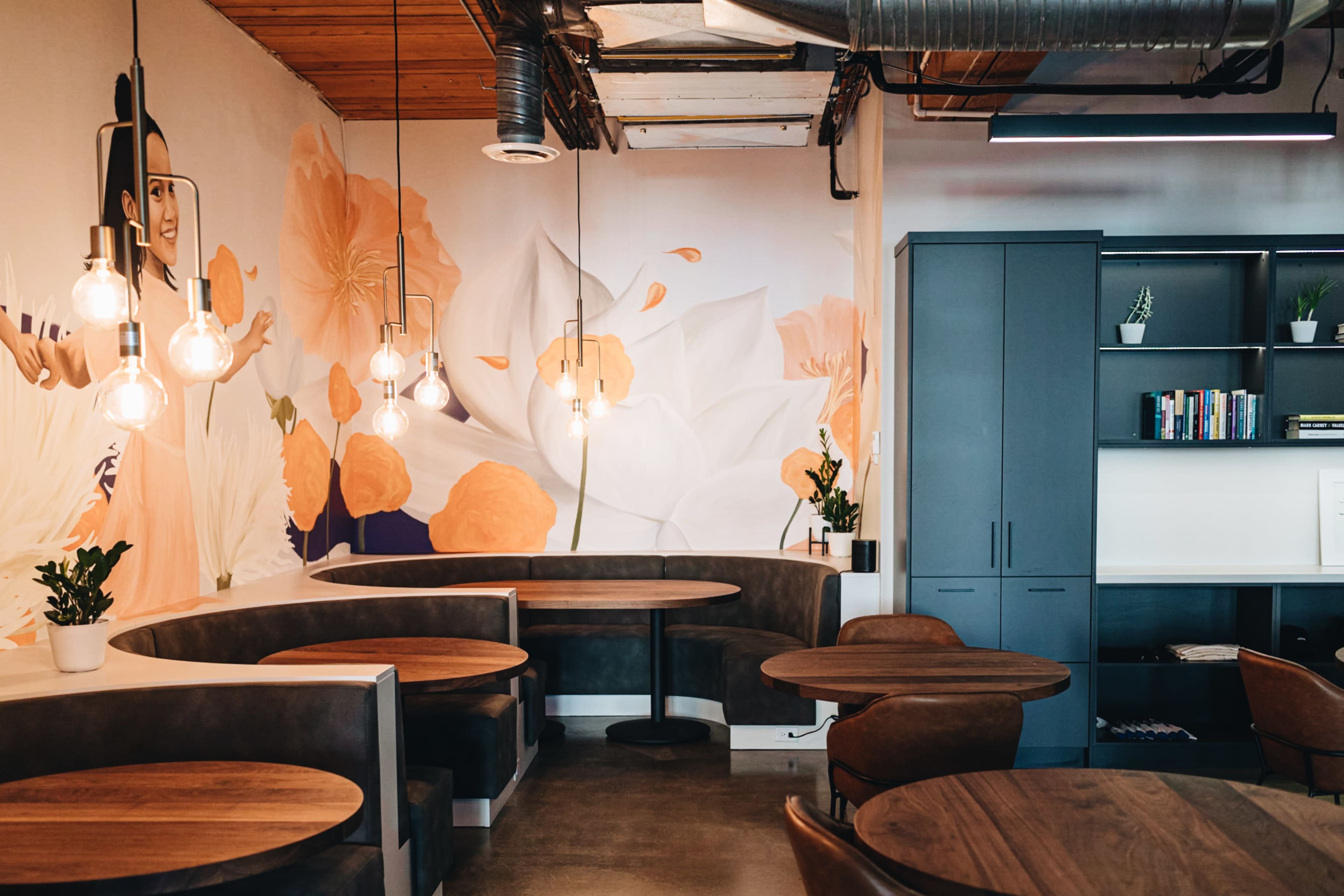 The image shows a modern café interior featuring round wooden tables, cozy booths, and a wall adorned with a floral mural.